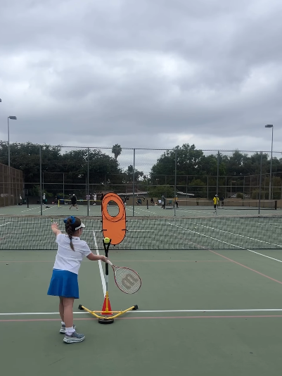 Child practicing tennis on an outdoor court with a tennis ball machine