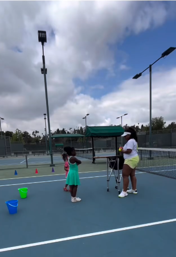 Two young girls and an adult woman playing tennis on an outdoor court with tennis balls, cones, and buckets, under a partly cloudy sky.
