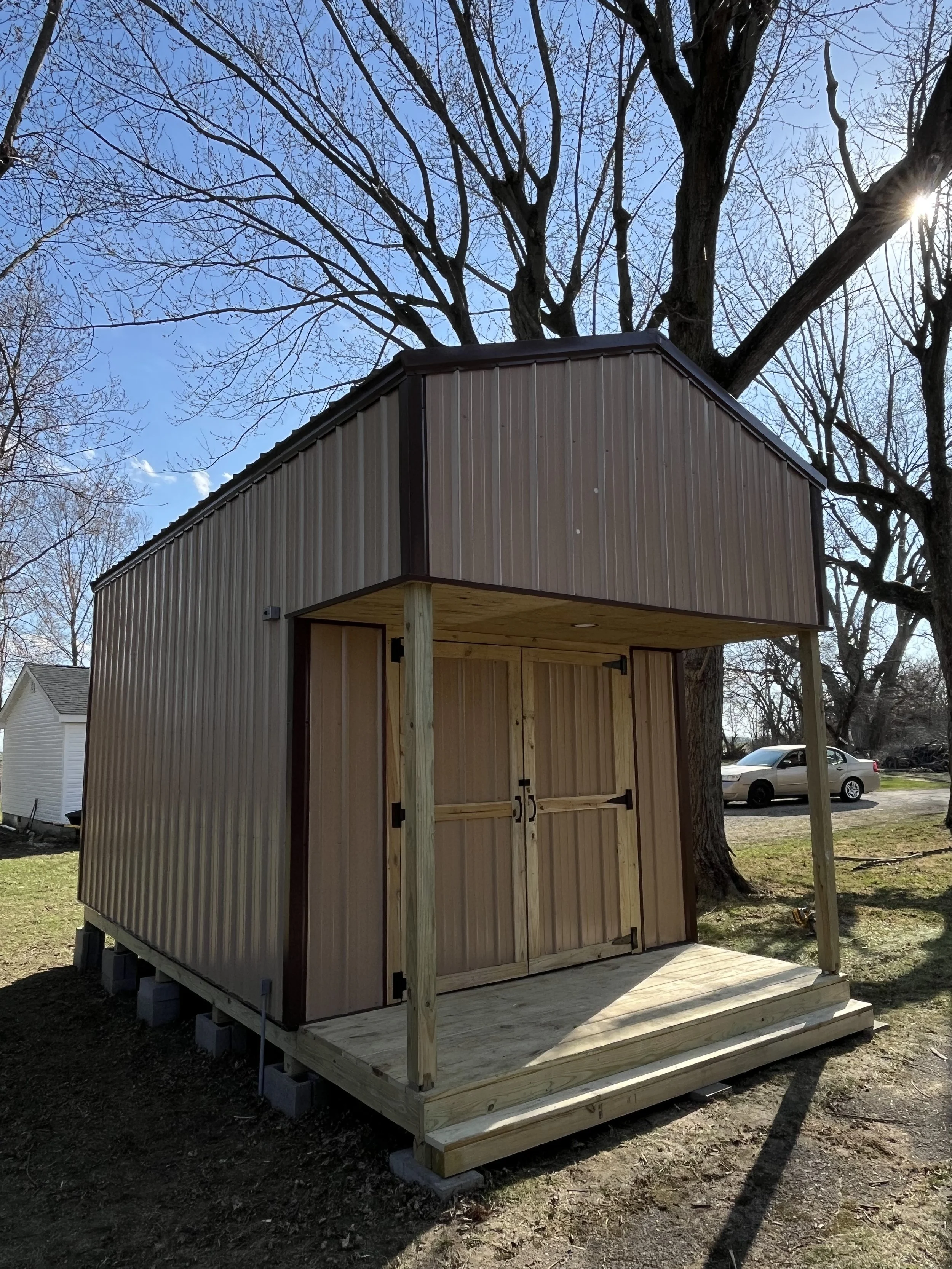 Small wooden structure under construction with vertical metal siding, opening for a door, supported by wooden posts, on a raised platform with steps, near trees and parked cars, on a sunny day.