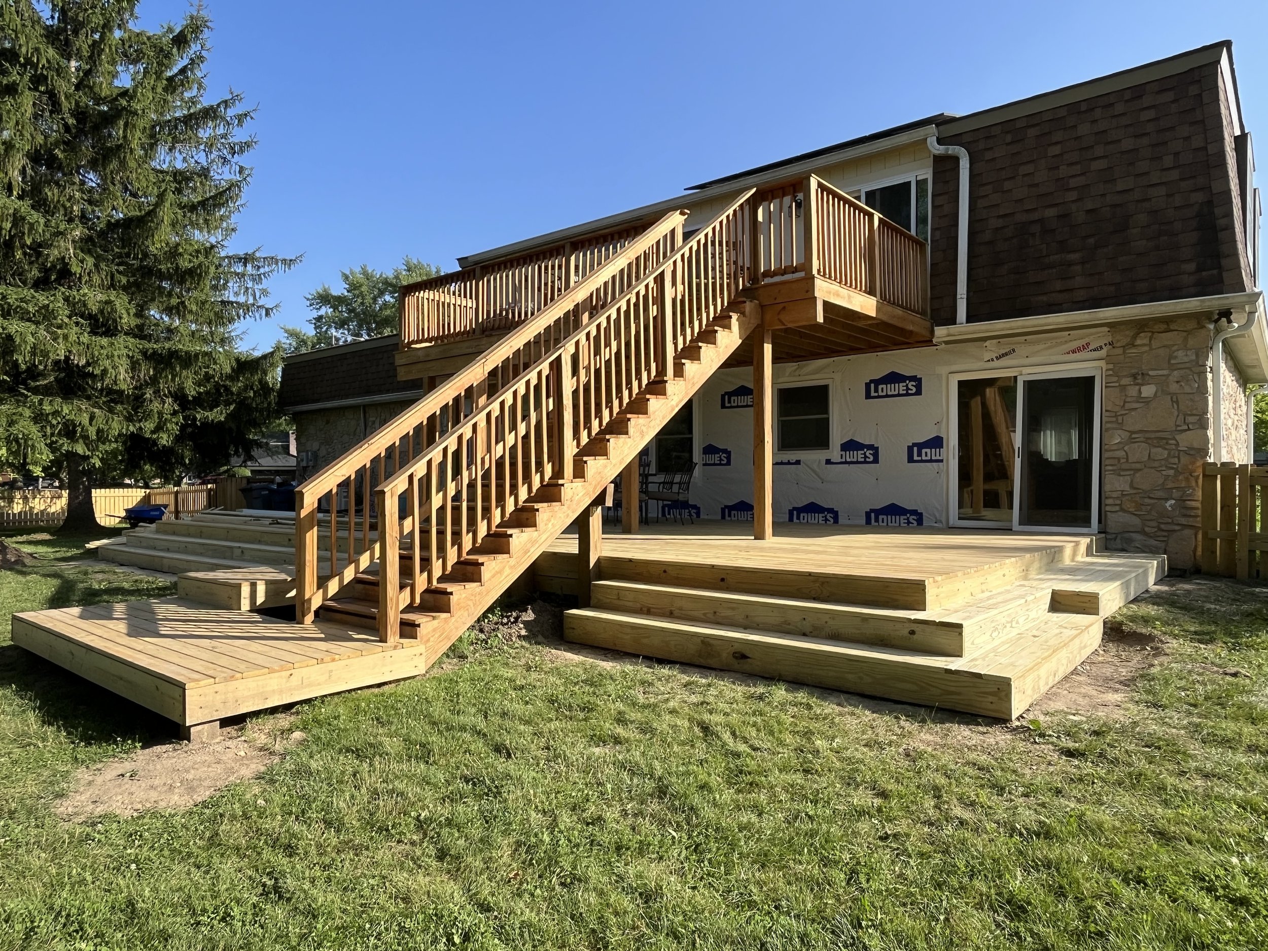 Newly constructed wooden deck with stairs attached to a house under renovation, with Lowe's signs on the house exterior, and a backdrop of a tree, grass, and a clear blue sky.