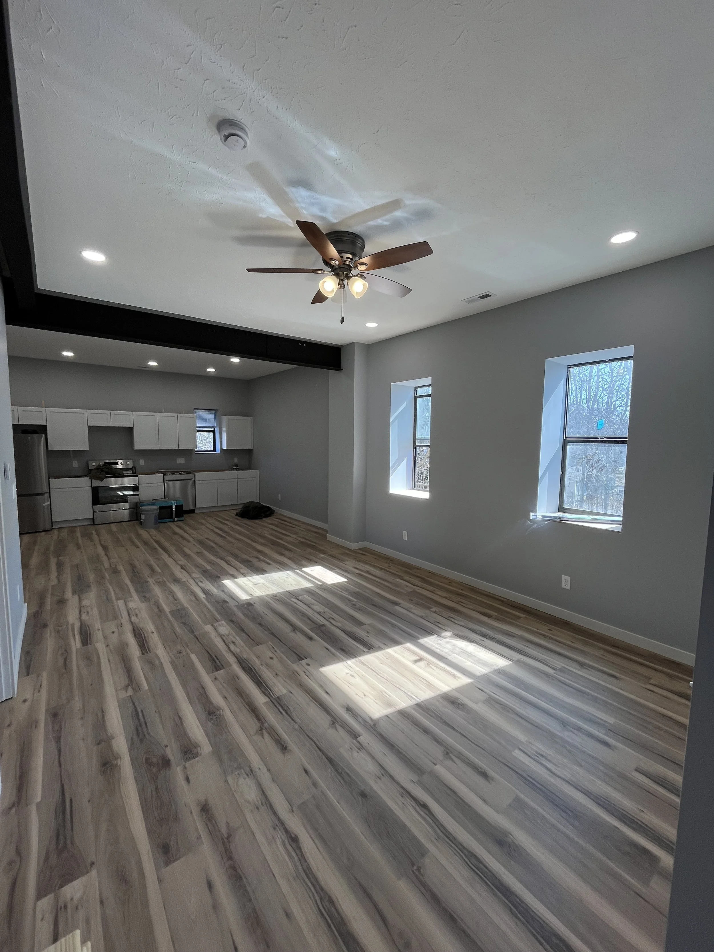 A spacious living area with light gray walls, wooden flooring, and windows letting in natural light. There is a ceiling fan with lights and a kitchen visible in the background.