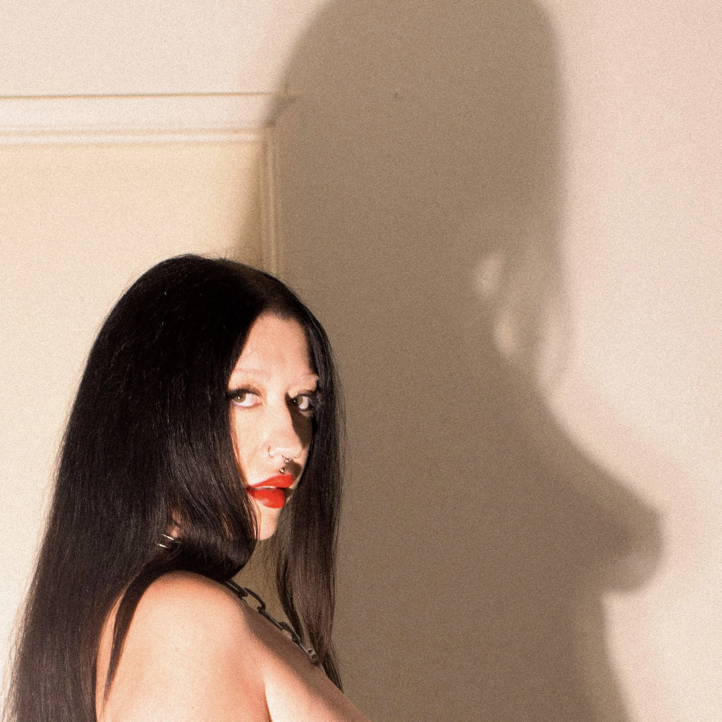 A woman with long dark hair, makeup, and a nose ring looks over her shoulder. She is wearing a black top with thin straps, and the background is a plain light-colored wall.