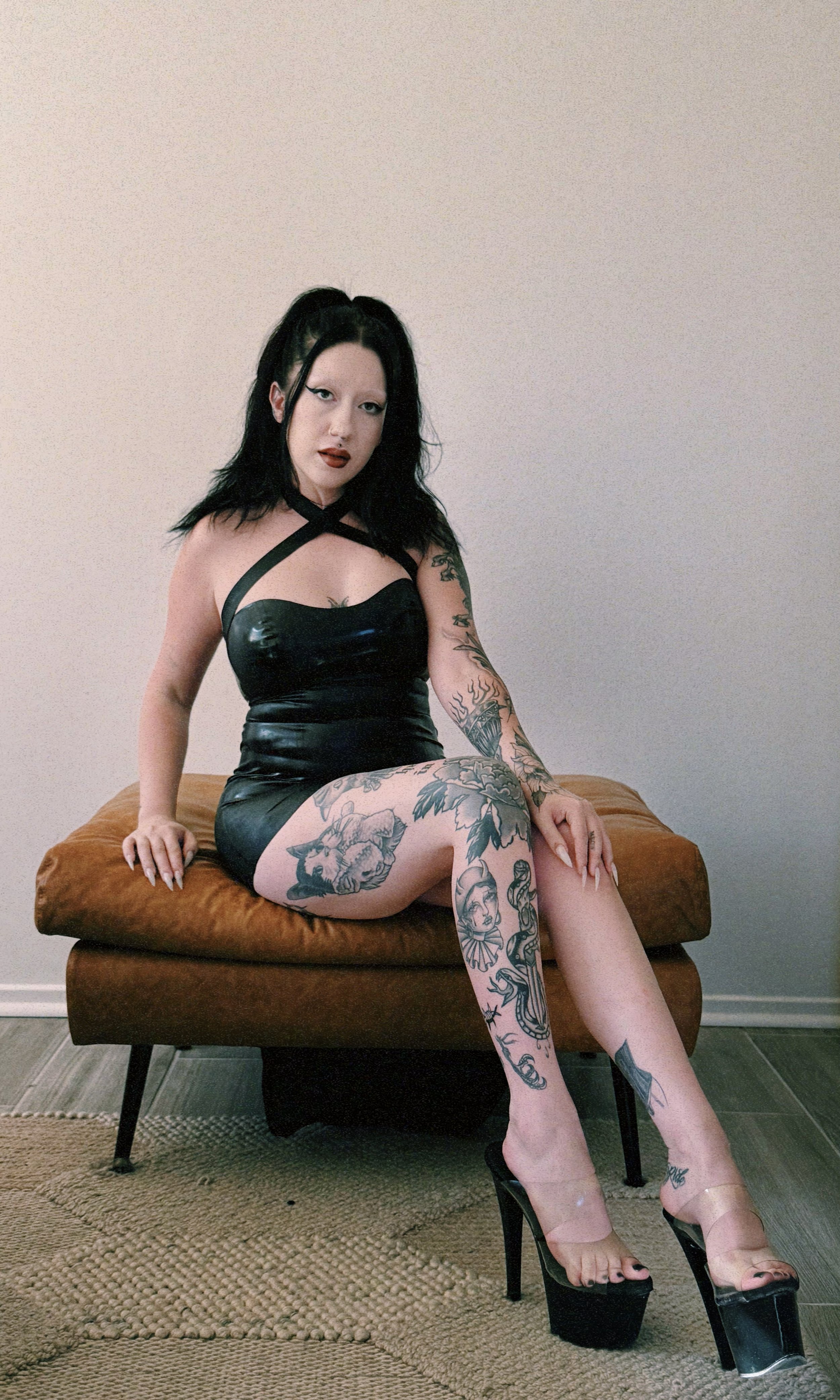 A woman with black hair, tattoos, and makeup sitting on a brown pouf. She is wearing a black dress with straps and black high-heeled platform shoes. The background is a plain wall and a rug is visible on the floor.