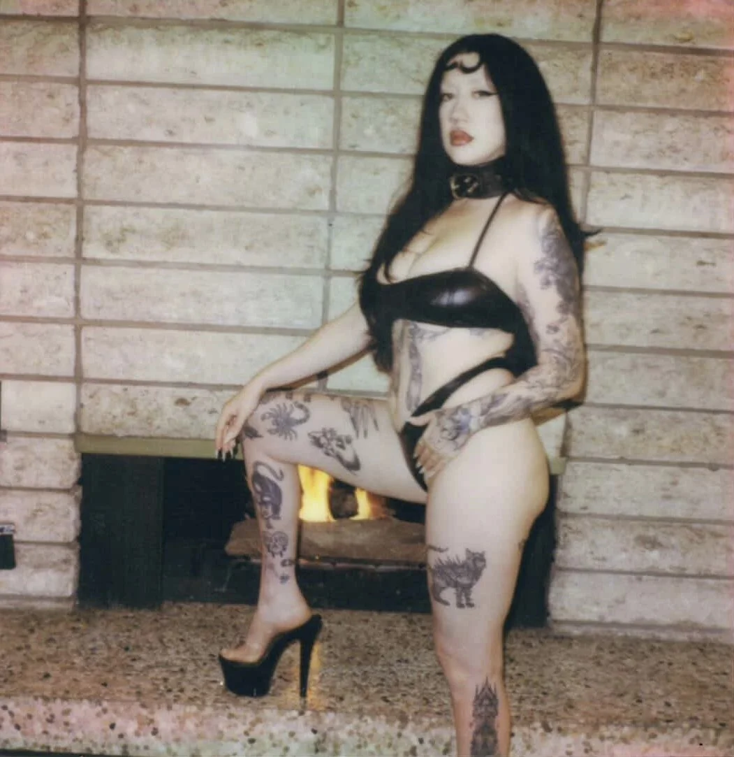 A woman with tattoos, dark hair, and makeup, wearing black lingerie and high heels, poses in front of a brick wall and a fireplace.