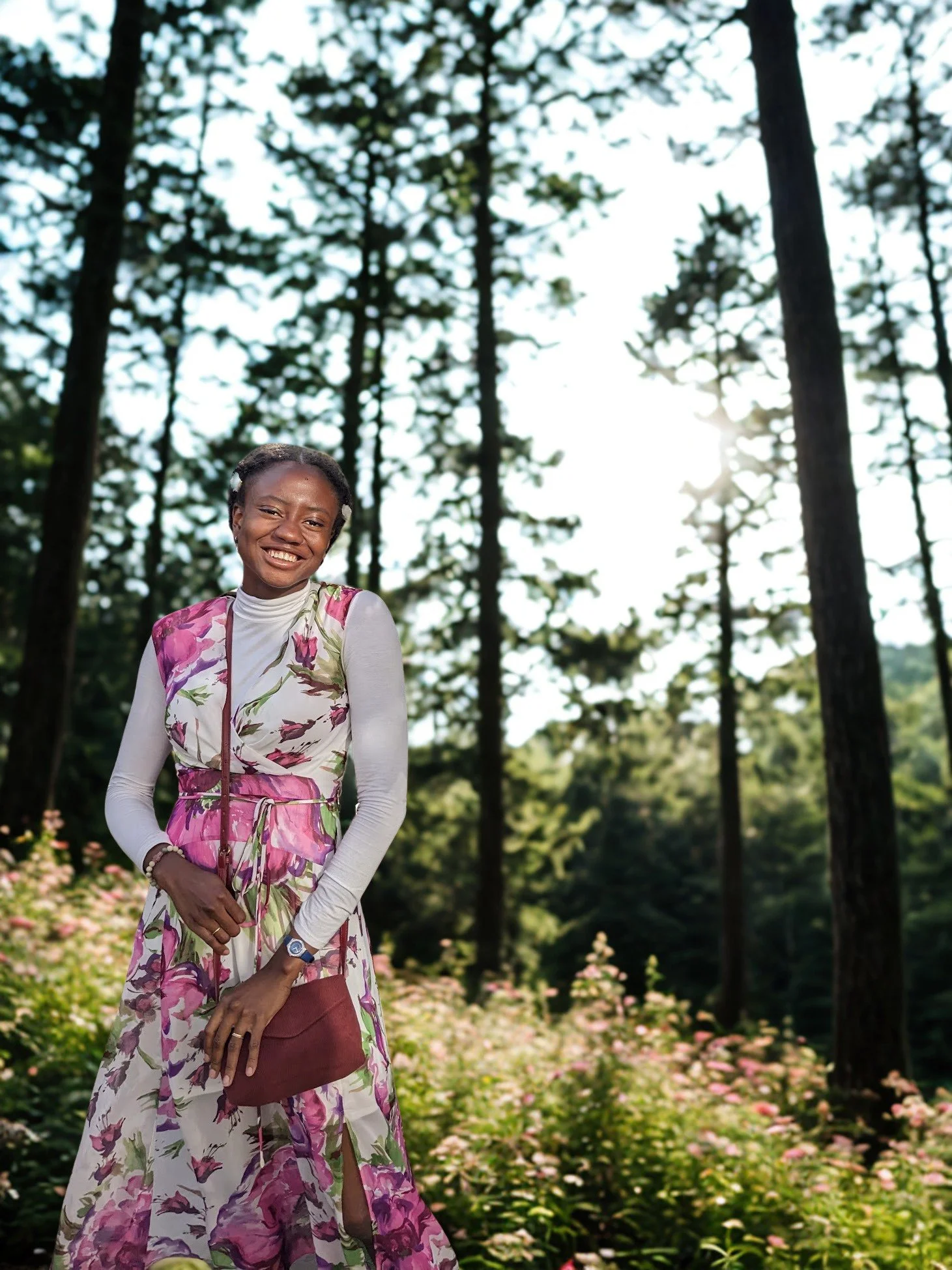 Victoria with a big smile, wearing a floral kimono-style dress with a white turtleneck underneath, standing in a forest with tall trees and sunlight shining through the branches, holding a maroon purse.