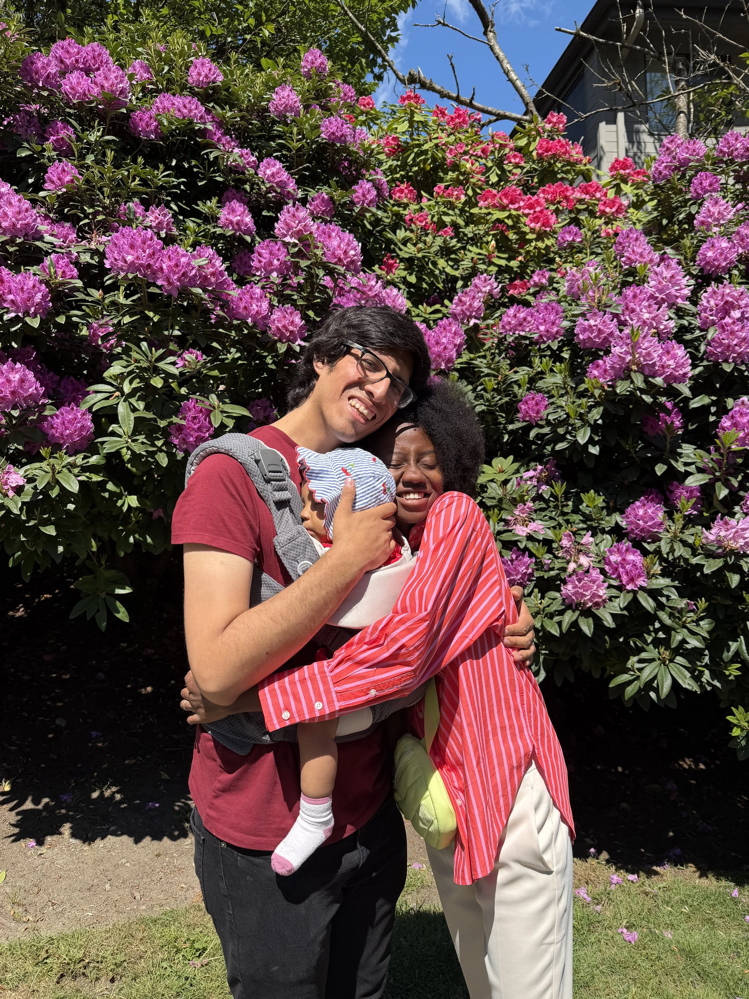 A family hugging in front of pink and purple rhododendron flowers, with a house and blue sky in the background.