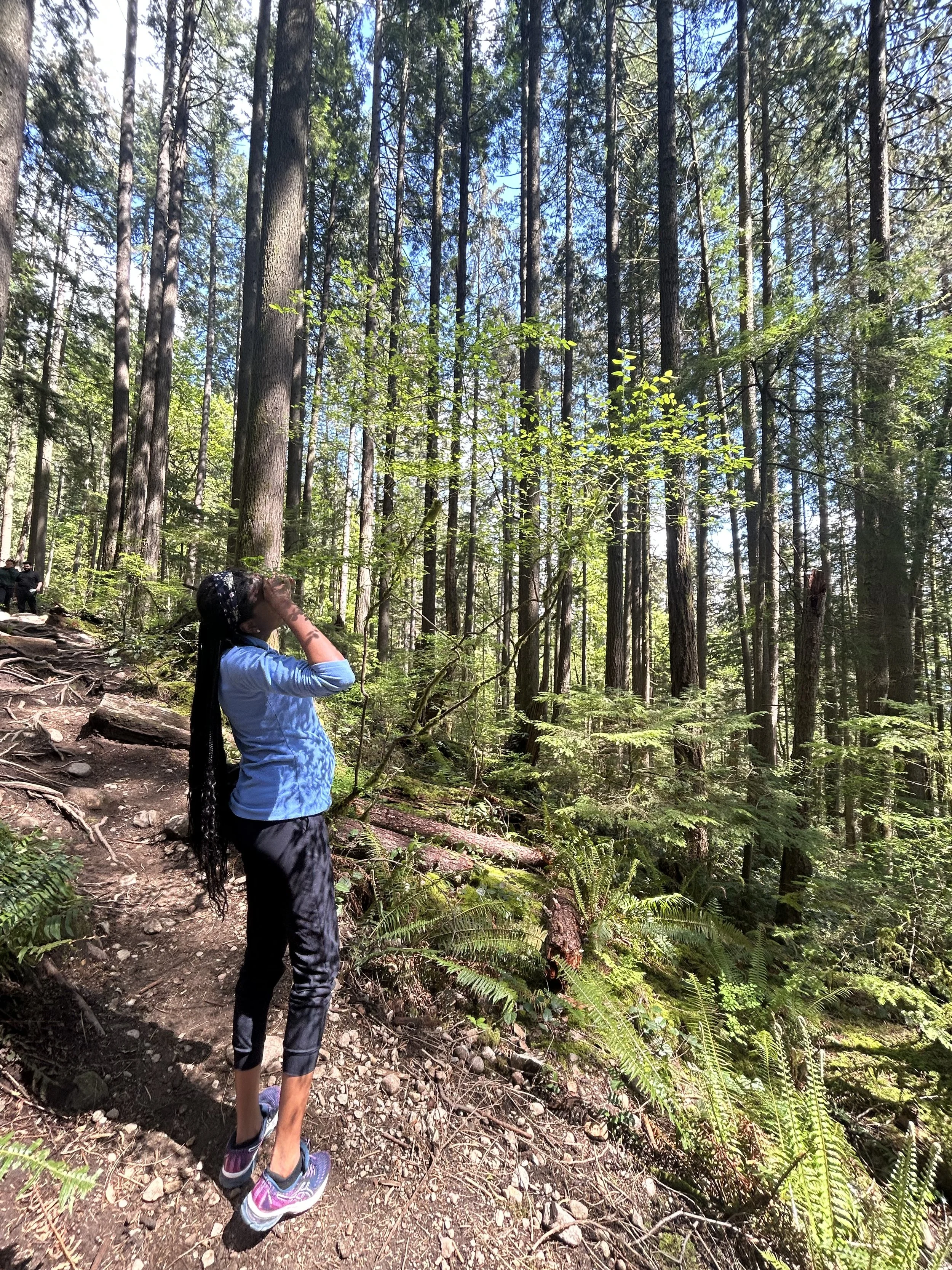 A person with long black hair, wearing a black headband, blue long-sleeve shirt, black pants, and colorful running shoes, standing on a dirt trail in a dense forest with tall trees and lush green foliage, looking upwards.