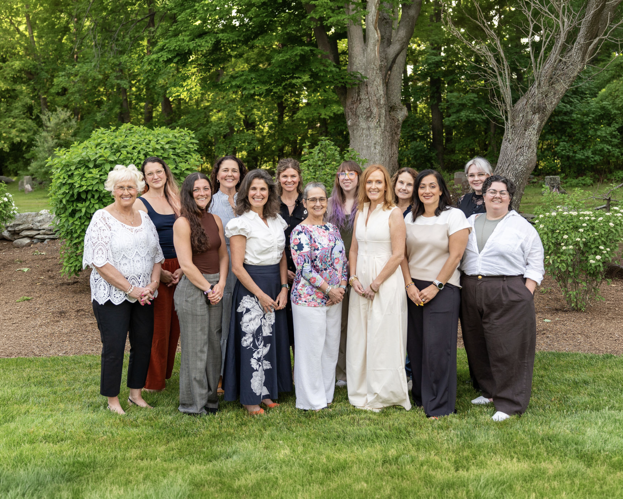 A group of women stand on green grass in front of trees.