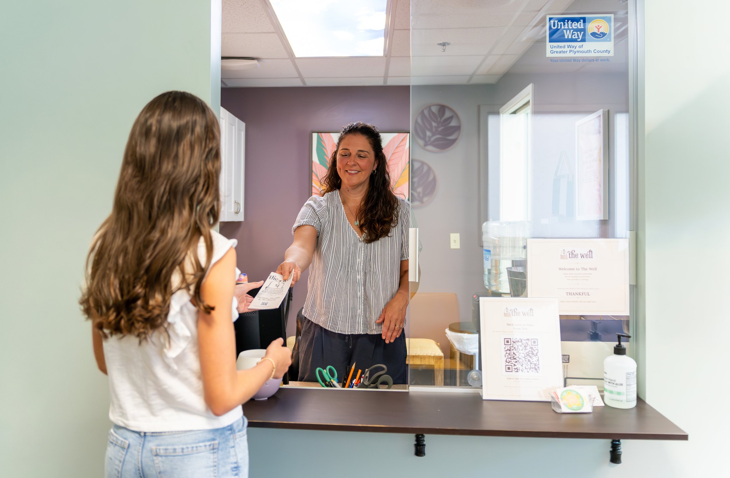 A woman behinds a desk stands to hand a young girl a pamphlet.