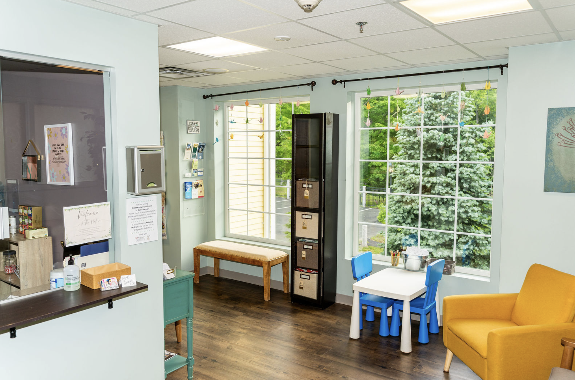 A room with blue walls has waiting chairs, a table with coloring supplies, and large windows.