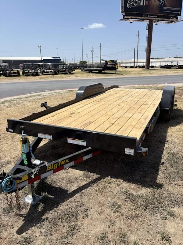 A flatbed trailer with a wooden deck parked on dirt beside a paved road, with trailers and a billboard in the background.