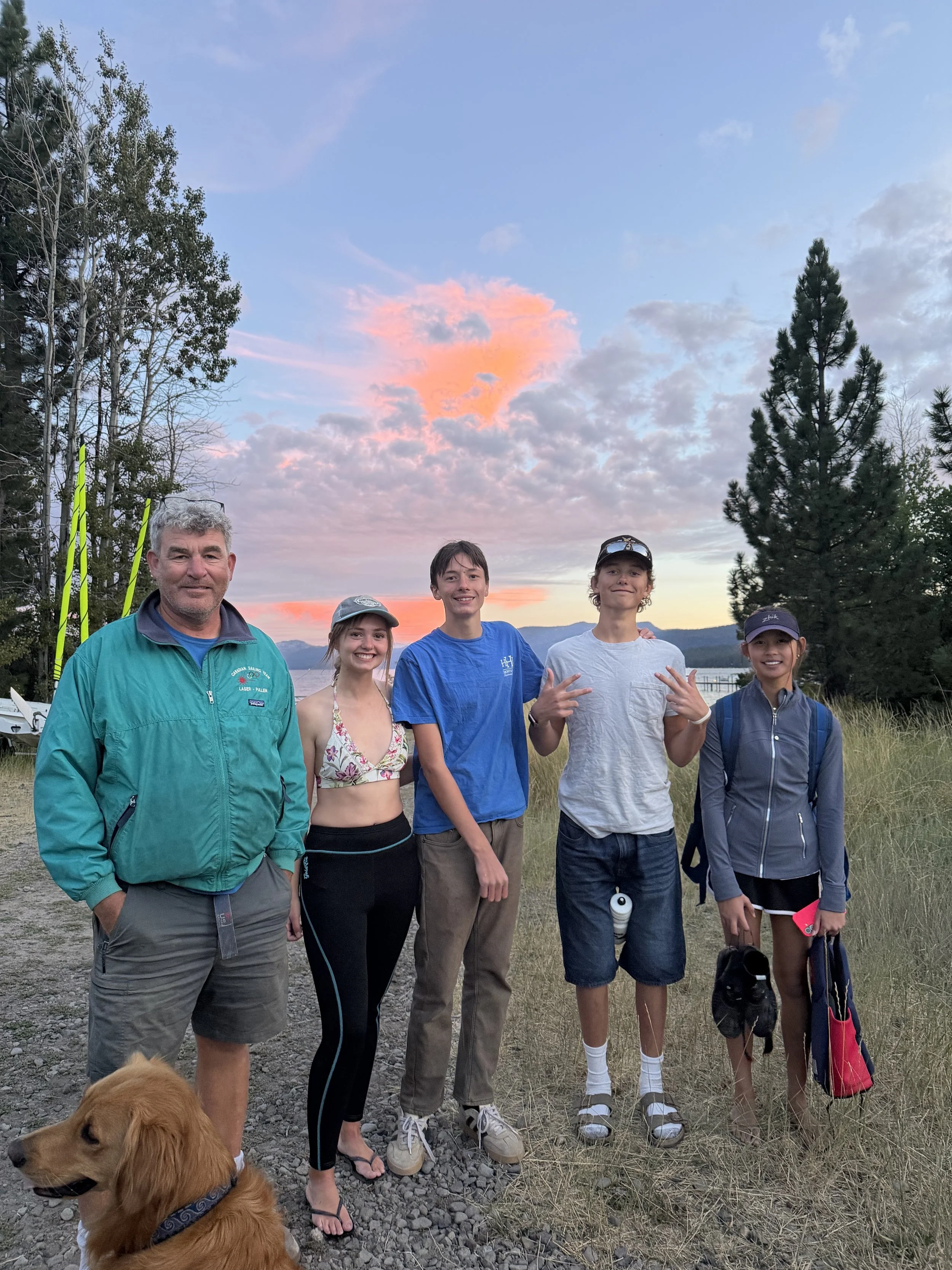 Group of six people, including men and teenagers, standing outdoors on a grassy area near a lake during sunset, with tall trees and cloudy sky in the background. A golden retriever dog is in the foreground.