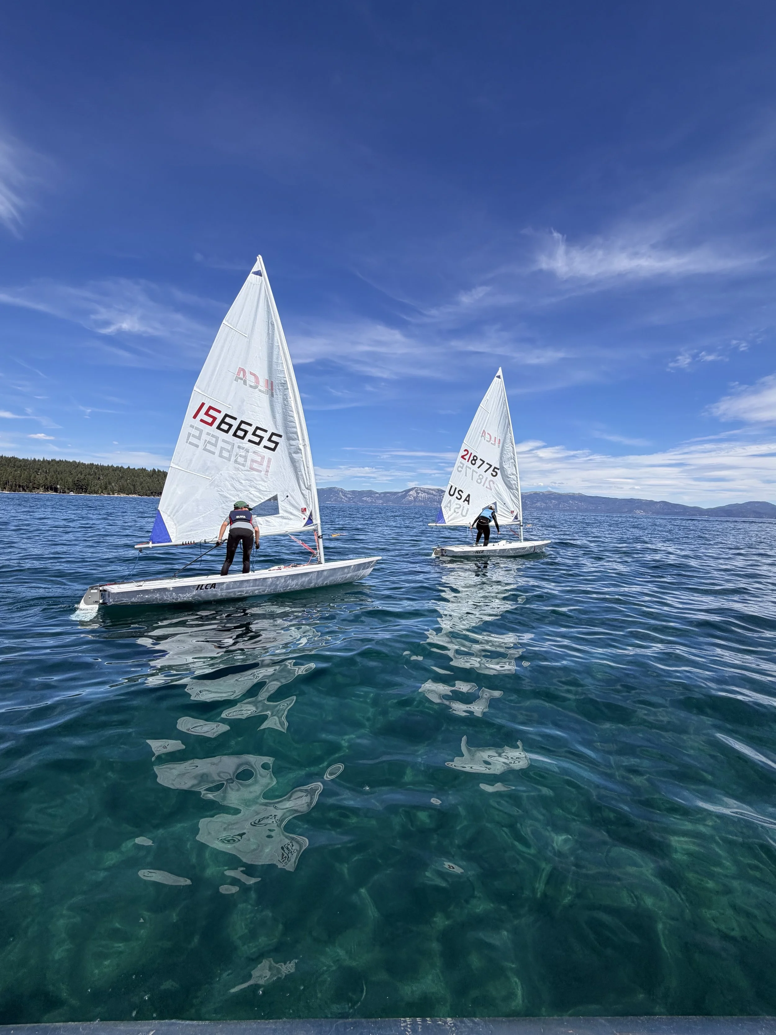 Two small sailboats with sailors on a large body of water, with mountains and a clear blue sky in the background.