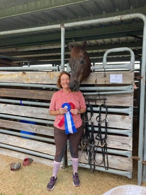 A woman holding a blue and red rosette prize ribbon standing next to a horse in a stable.