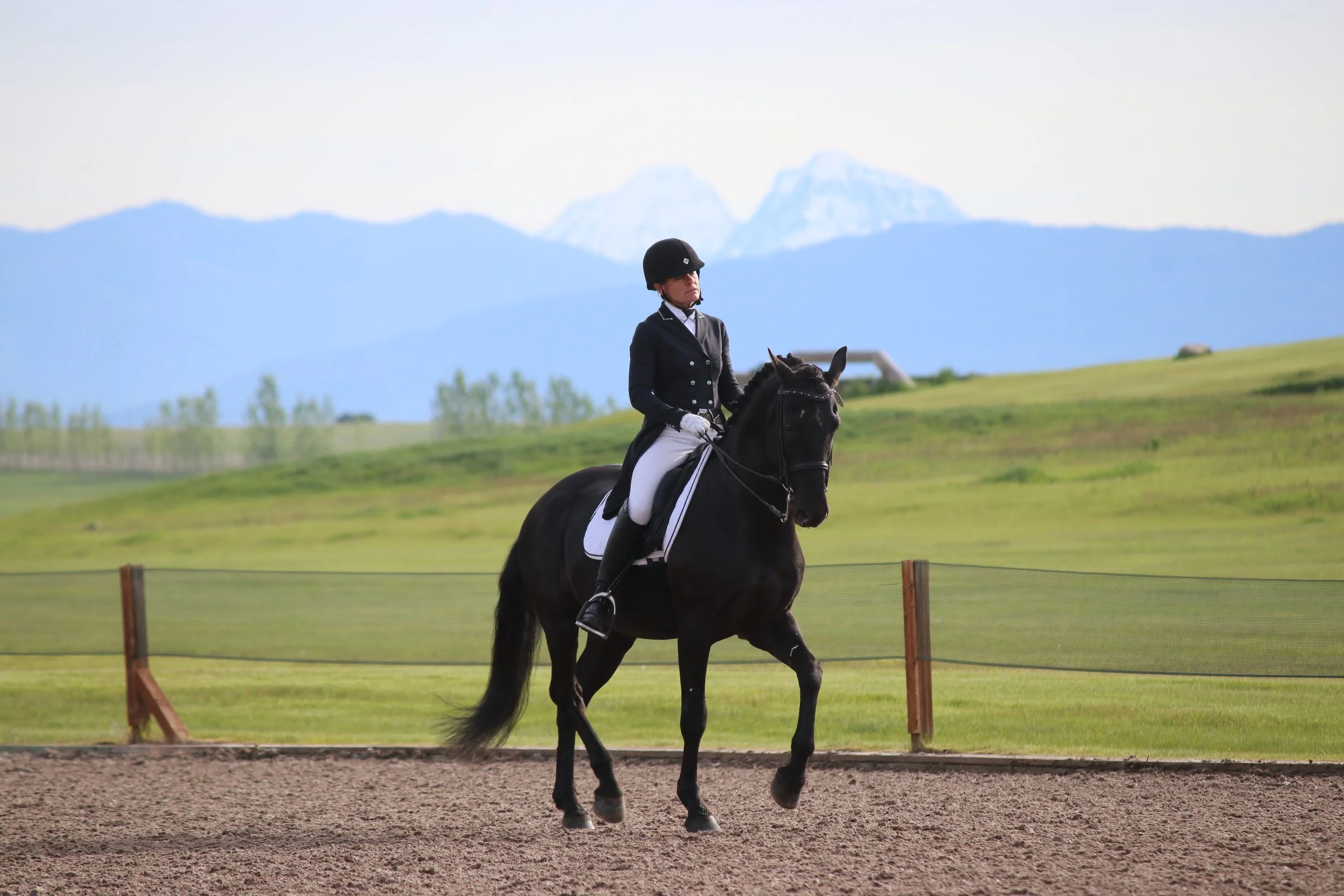 A person dressed in equestrian attire riding a black horse in an outdoor riding arena with a mountain range and green fields in the background.