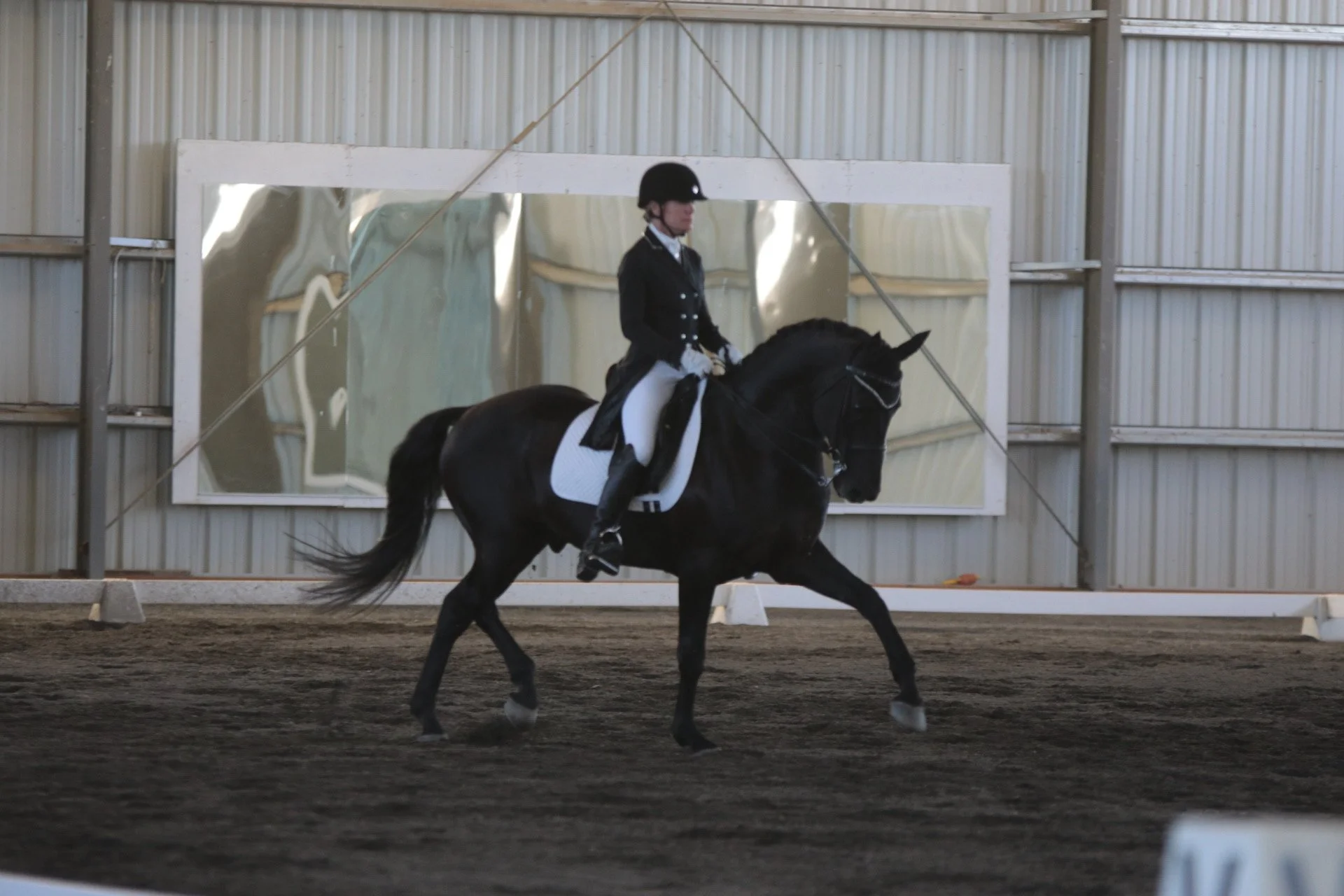 A rider dressed in formal equestrian attire, including a black helmet and jacket, is riding a black horse inside an indoor riding arena. A large mirror and metal siding are visible on the arena's wall.