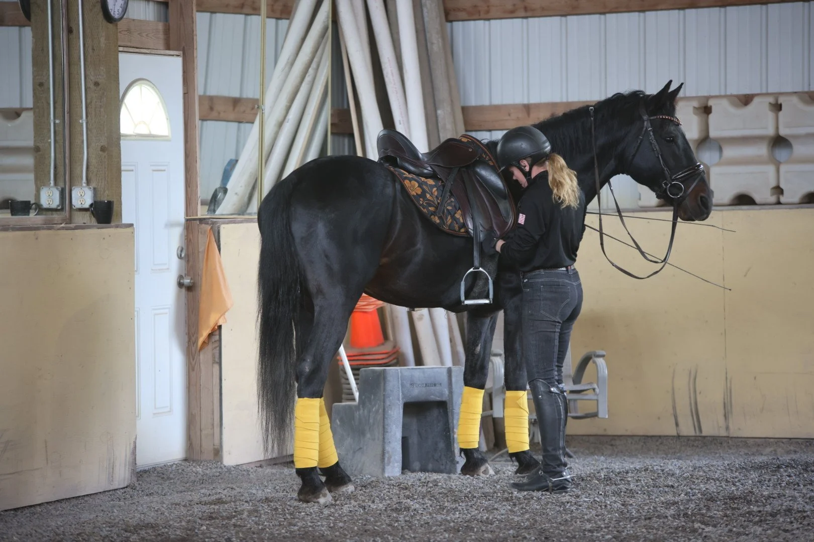 A person in black riding gear helping a dark brown horse with yellow bandages on its legs to get ready for riding inside a stable or riding arena.