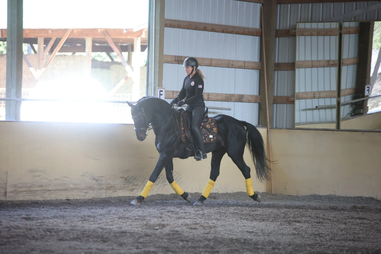 A person riding a black horse in an indoor riding arena, wearing a helmet, black riding outfit, and yellow leg wraps, with a large mirror on the right side.
