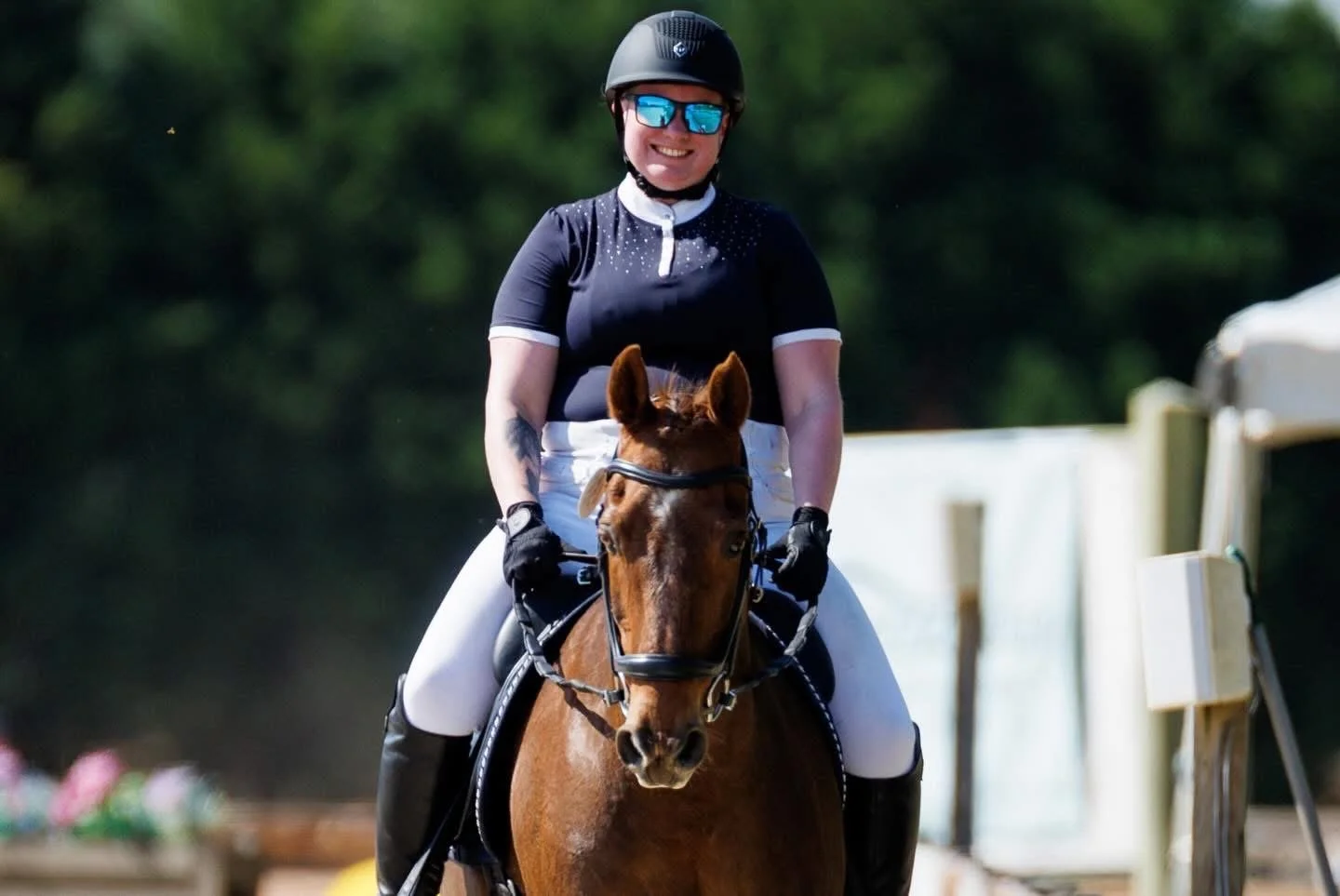 A woman smiling while riding a brown horse, wearing a black helmet, sunglasses, and equestrian attire, outdoors with a blurred green background.