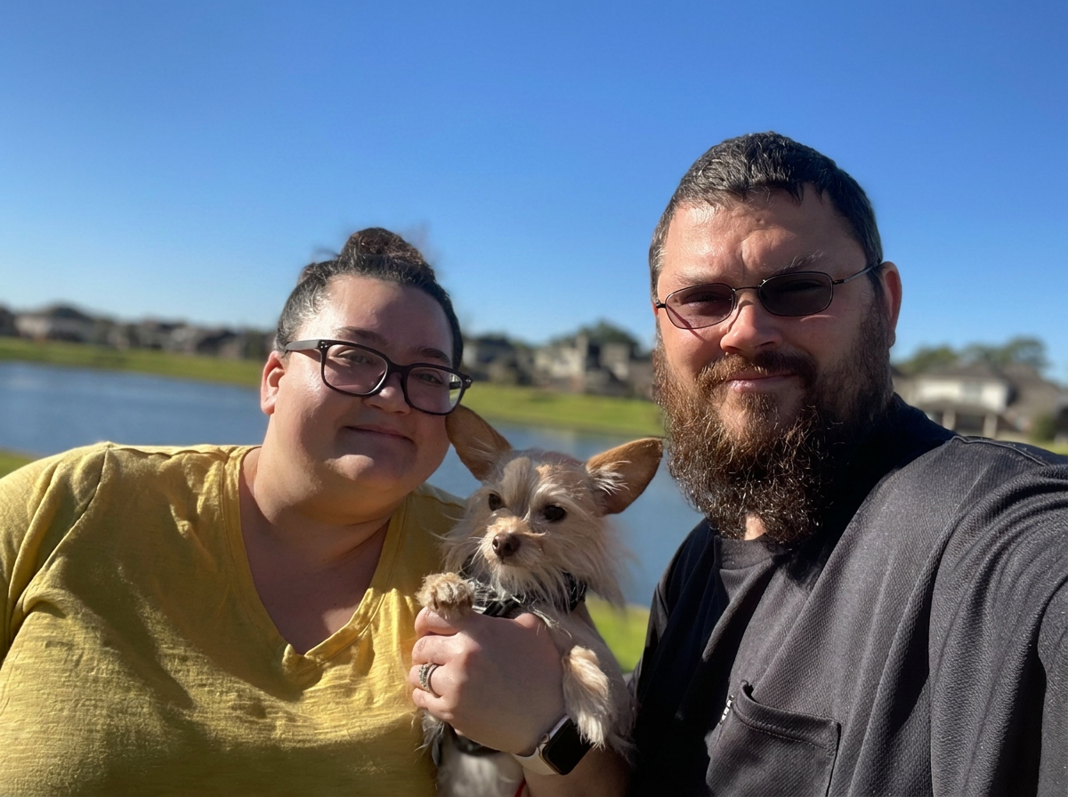 A man and woman holding a small dog outdoors near a body of water with houses in the background under a clear blue sky.