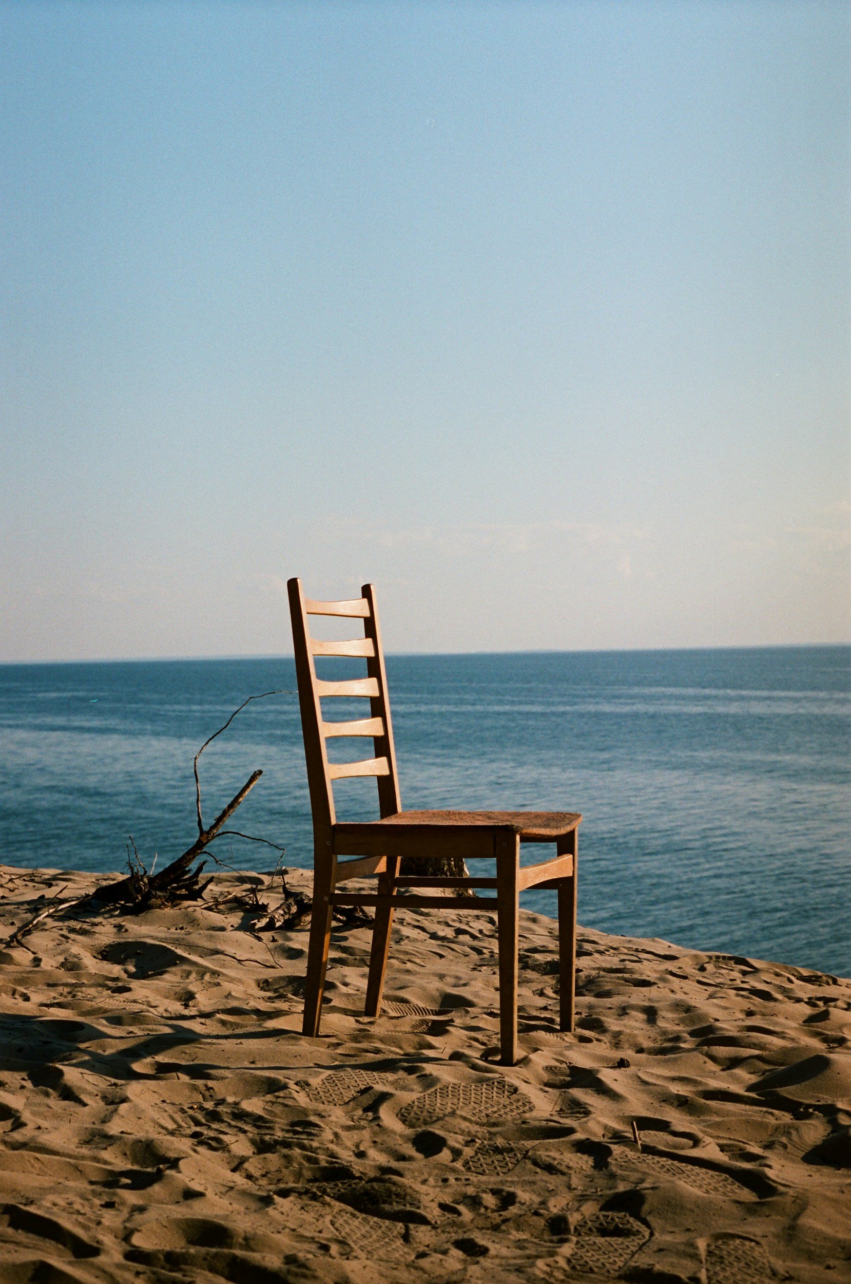 A wooden chair sitting on sandy beach with sea and clear sky in the background.