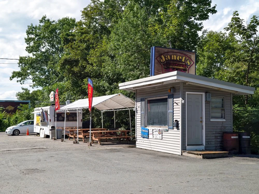 A small roadside hot dog truck named Janet's Chili Dogs with an outdoor seating area under a canopy, set against a background of trees and a partly cloudy sky.