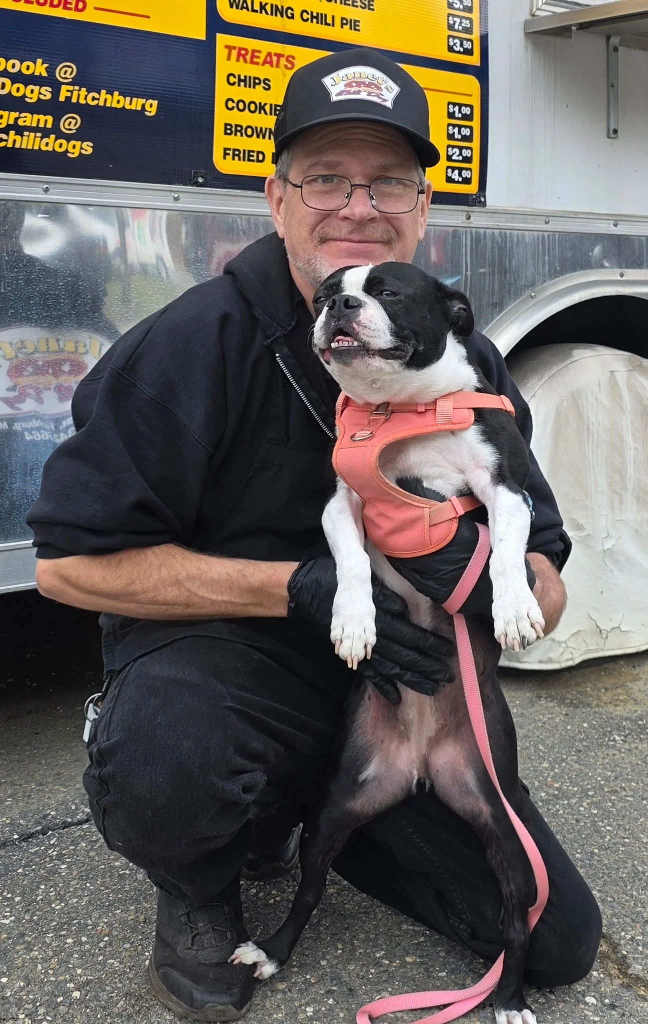 A man with glasses, a gray beard, wearing a black at, black jacket, and gloves is squatting next to a black and white dog with a pink harness. The man is smiling and holding the dog, which appears to be a Boston Terrier or similar breed. They are outside at a food truck with a yellow menu board in the background.