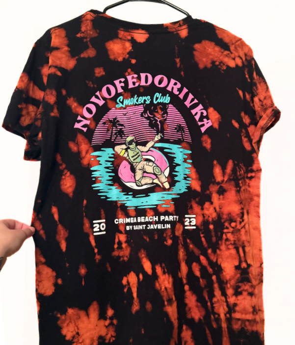 Tie-dye T-shirt with a graphic and text promoting a beach party. The shirt has a black and orange pattern. The graphic features a person in a boat with a pink inner tube, on water with palm trees and an animal, possibly a lion, in the background. The text includes "NOVOFEDOROVKEV" at the top, "Smokers Club," "Crimea Beach Party," and "by Saint Javelin," with the years 2022 and 2023.
