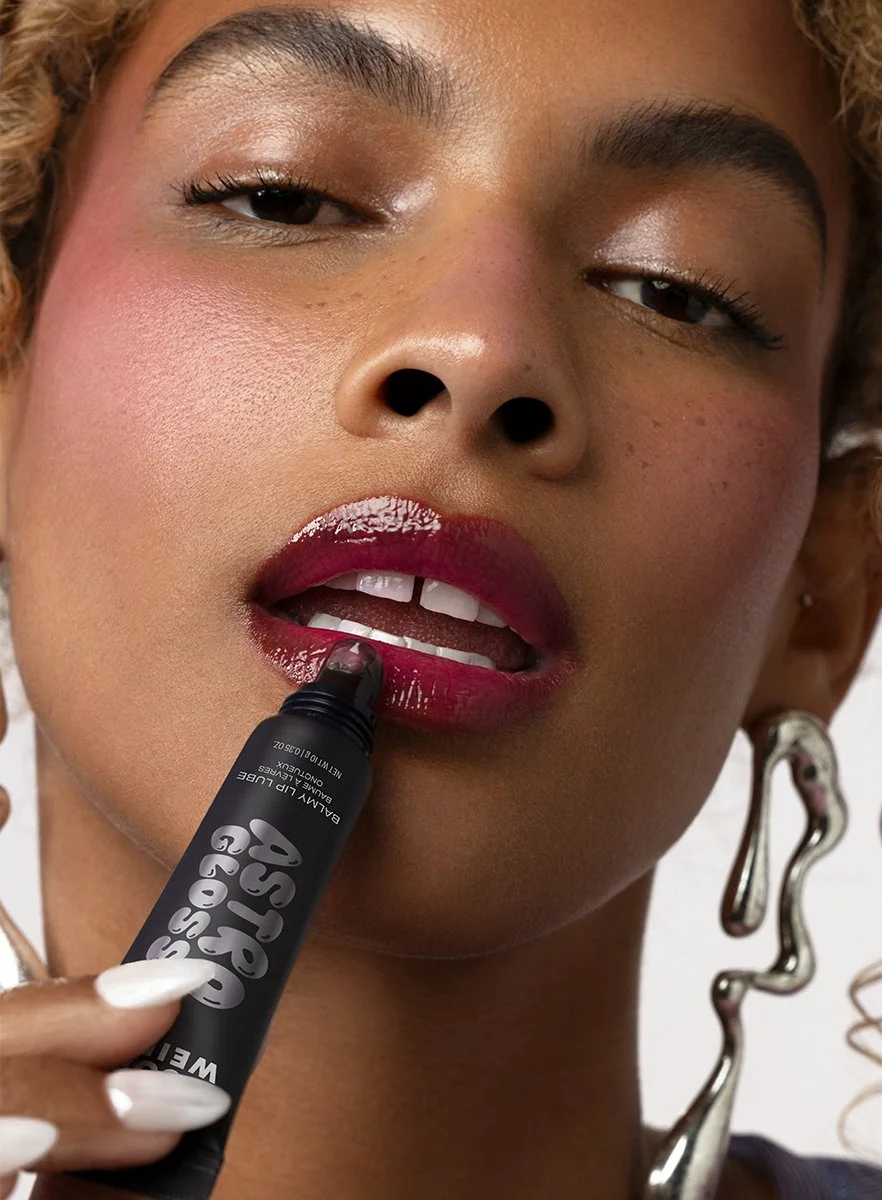 A woman with curly blonde hair applies red lipstick with a black marker to her lips, which are slightly parted.