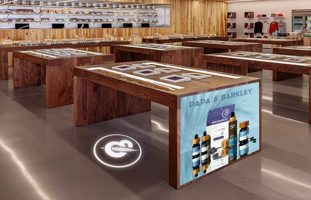Modern retail store with wooden display tables showcasing electronic devices, including tablets. A digital circular sign on the floor features a stylized arrow and an ear of wheat, with additional shelves and clothing in the background. Promotional s
