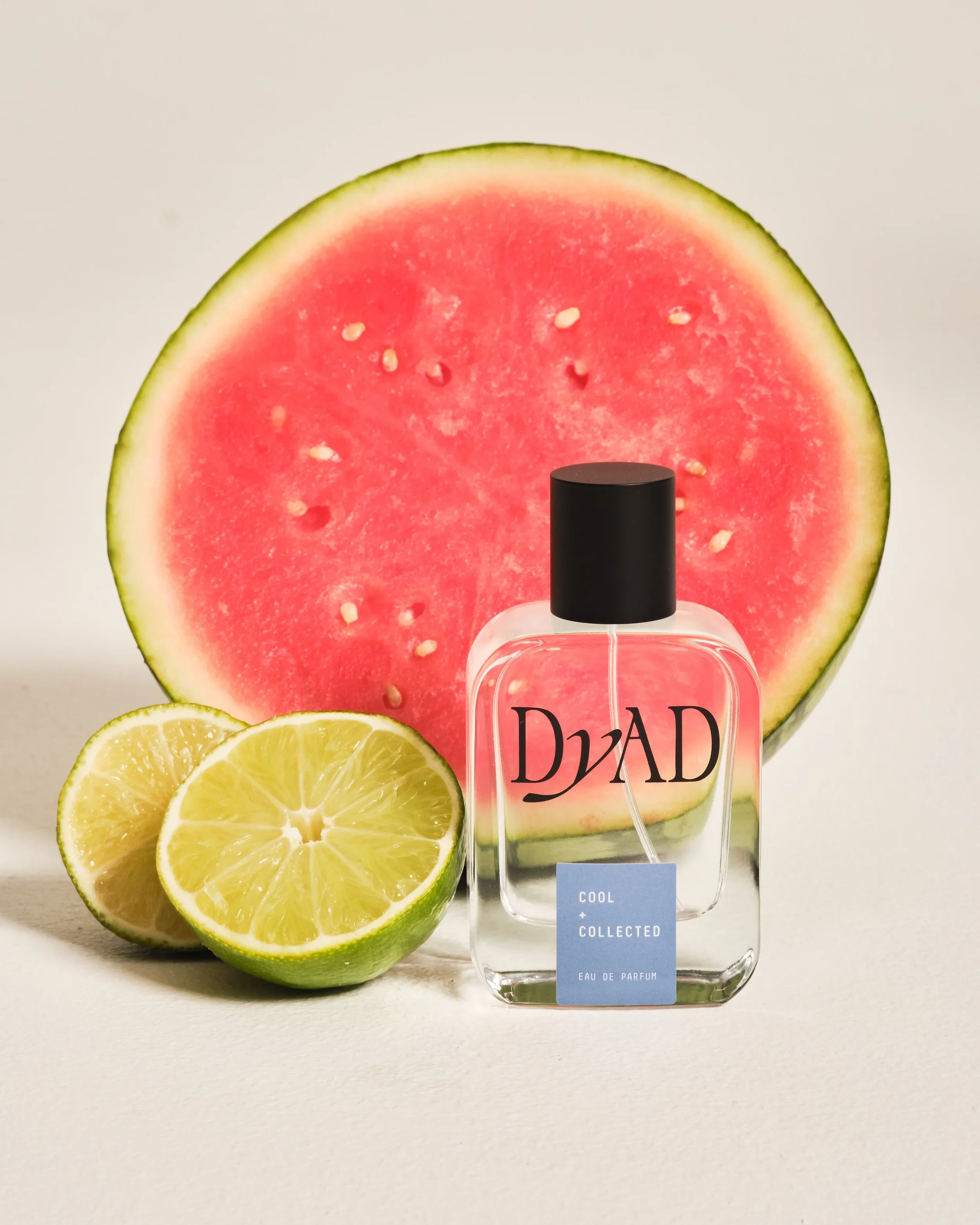 A close-up of a perfume bottle labeled 'D2AD' with slices of watermelon, lemon, and lime surrounding it on a white background.