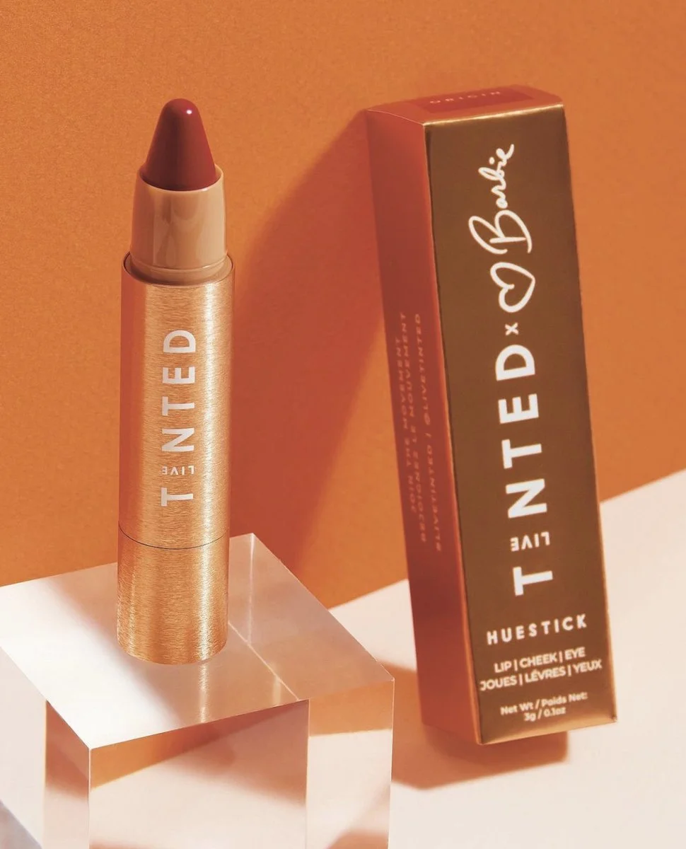 A tube of Tinted Love lipstick with a gold-colored finish, standing upright on a clear acrylic stand, next to its packaging box on an orange background.
