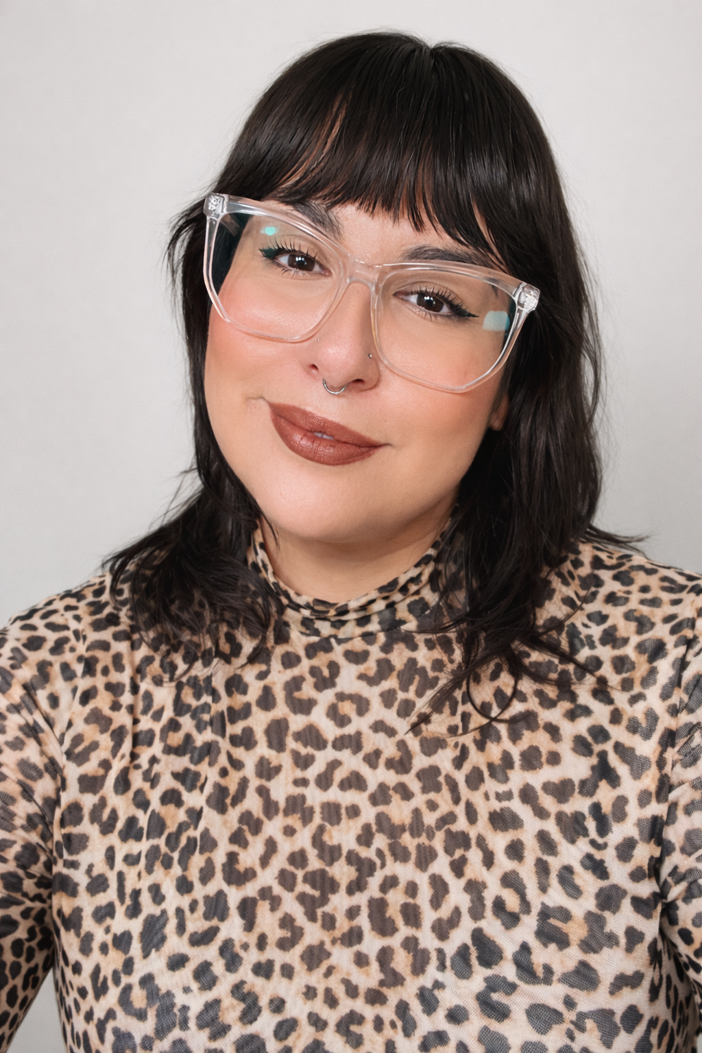 A woman with dark, wavy hair, wearing clear glasses, a septum piercing, and a leopard print top, smiling at the camera against a plain background.