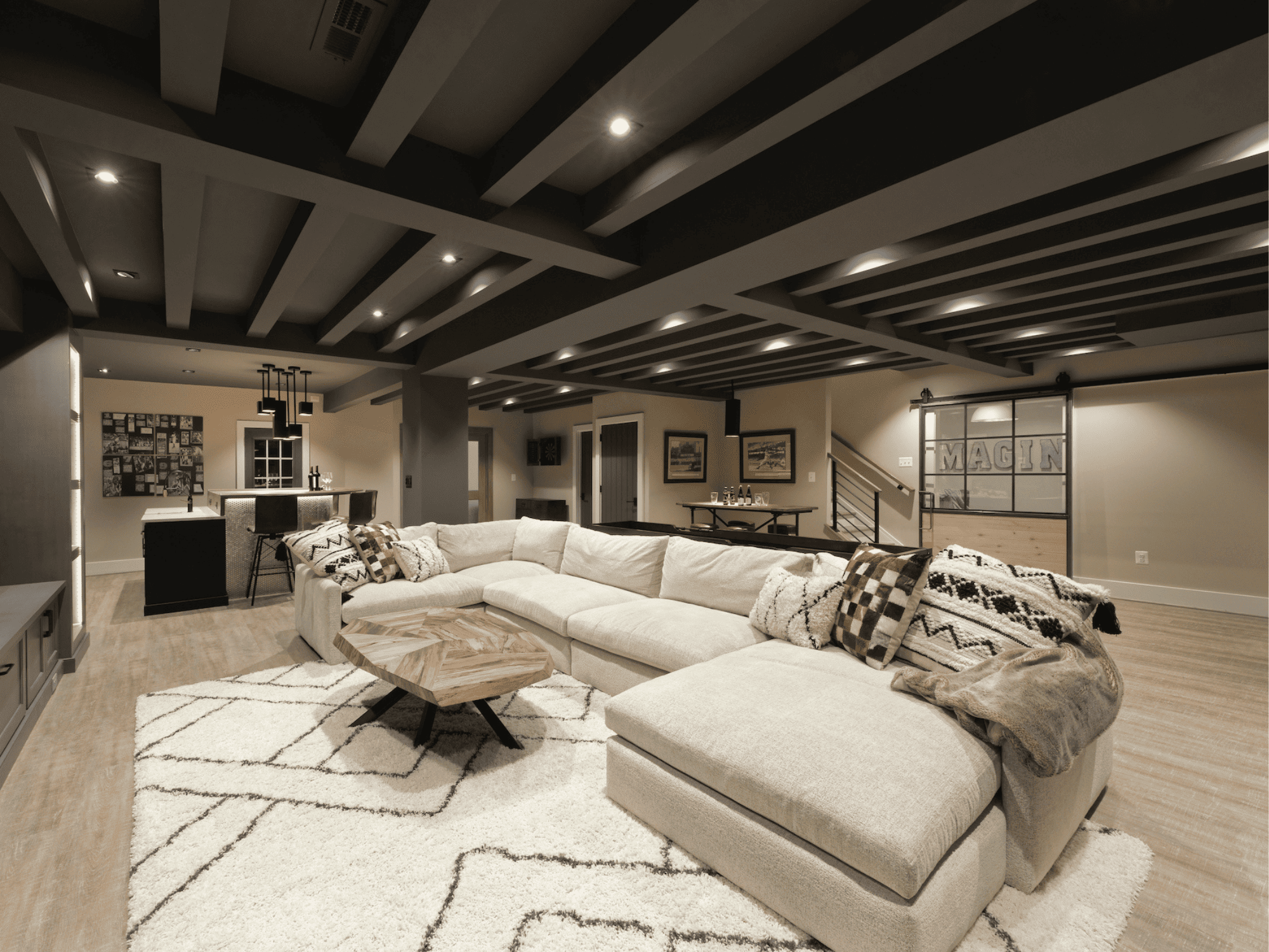 A spacious living room with a large cream sectional sofa decorated with various pillows, a patterned rug, and a modern geometric wood coffee table. The ceiling has black and gray wooden beams with recessed lighting, and the background includes a dining area with a dark table, pendant lights, and framed wall art.
