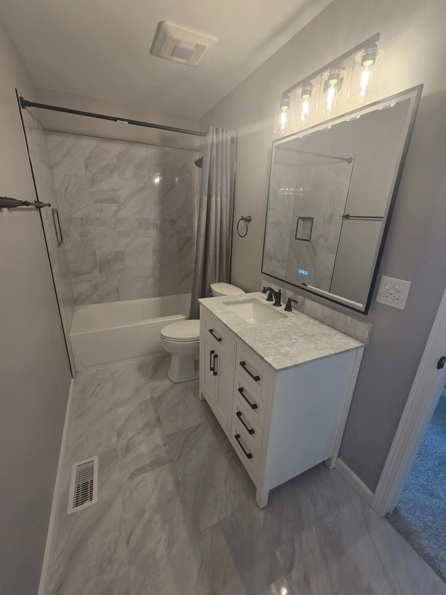 Bathroom with marble tiles, a bathtub with a shower curtain, a toilet, a white vanity with a sink, a large mirror, and a row of lights above the mirror.