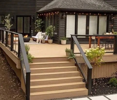 Outdoor wooden deck with stairs leading up to a black house decorated with string lights, potted plants, and outdoor furniture.