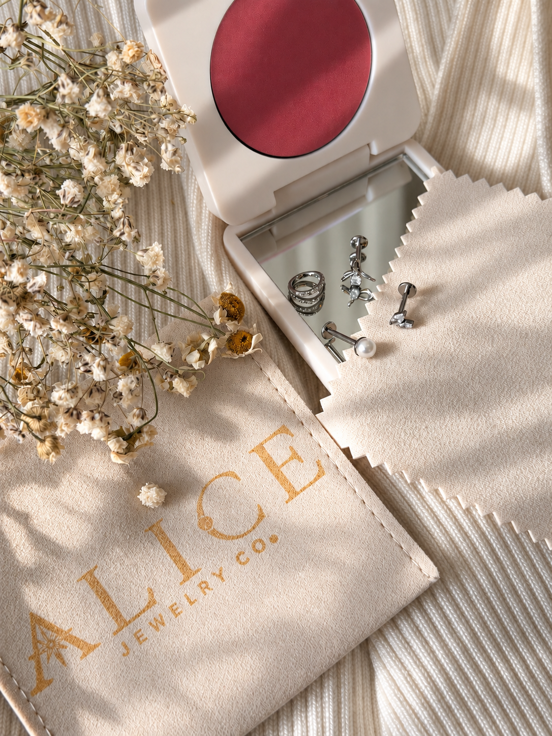 A jewelry display box with earrings and rings, a beige jewelry pouch with "ALTEJEWELRY CO." printed on it, a bouquet of dried flowers, and a compact mirror, all arranged on a textured cream-colored fabric surface.