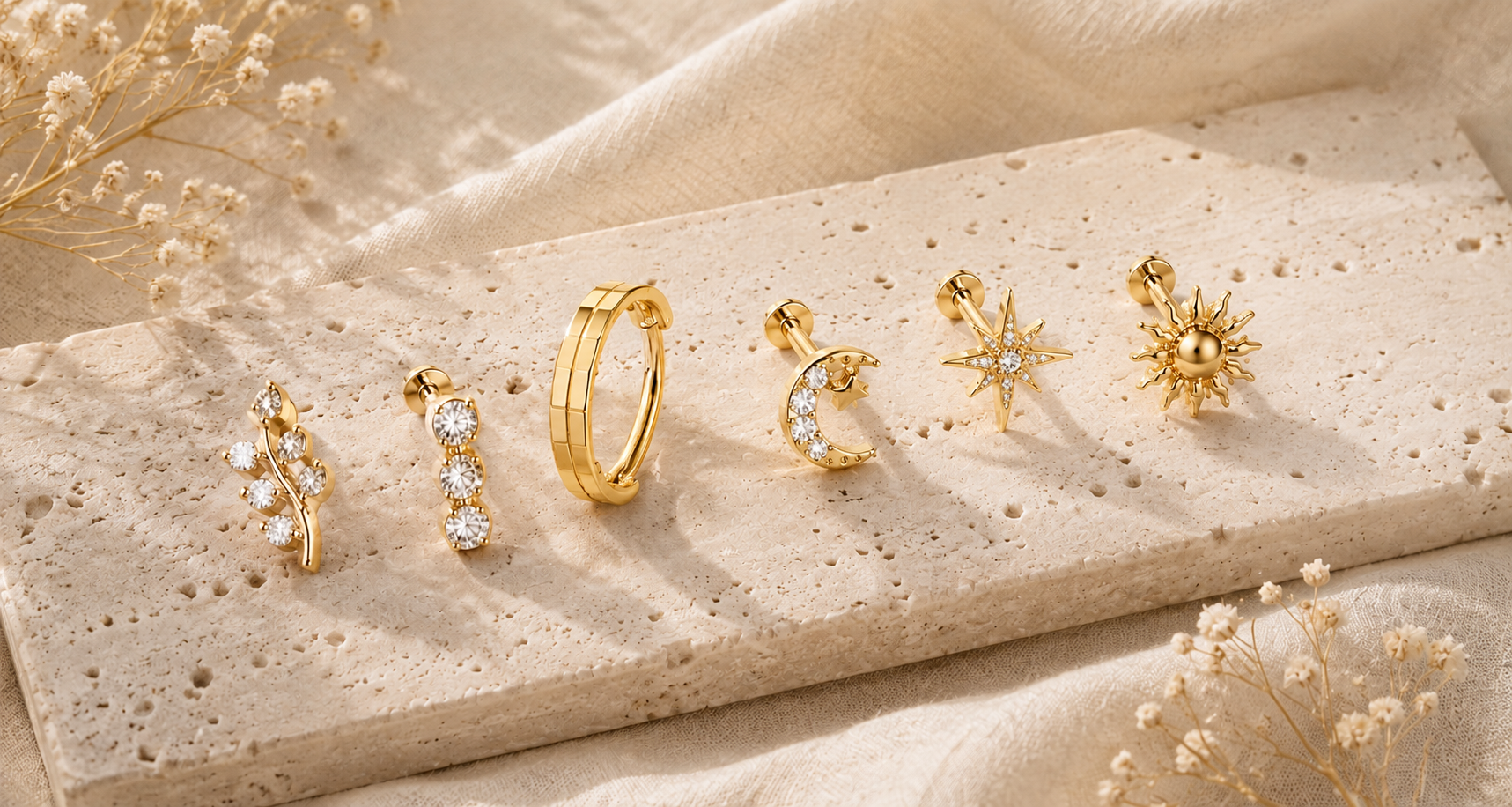 A display of six gold earrings with celestial and nature-inspired designs, set on a beige textured surface with dried flowers around.