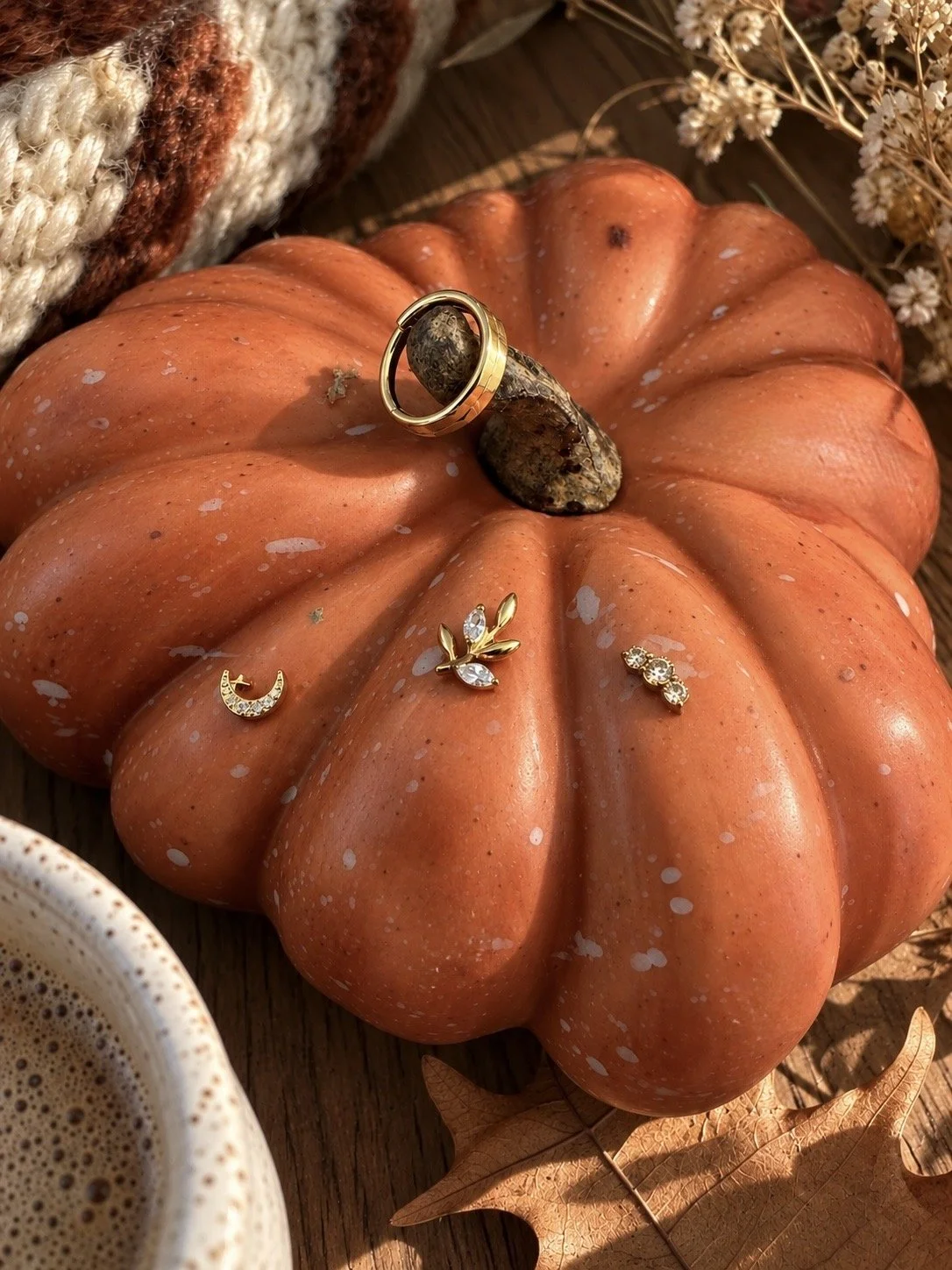 An orange pumpkin with jewelry placed on top, including a gold ring, earrings, and a crescent moon pendant. There are fall leaves, a cup of coffee, and dried flowers around the pumpkin on a wooden surface.
