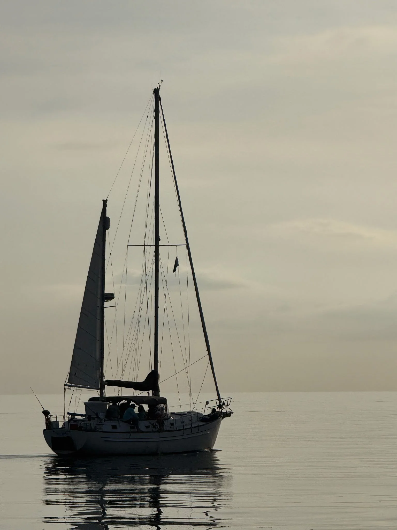 A sailboat with a tall mast and a partially rolled sail floats on calm water under a cloudy sky, with its reflection visible on the water's surface.
