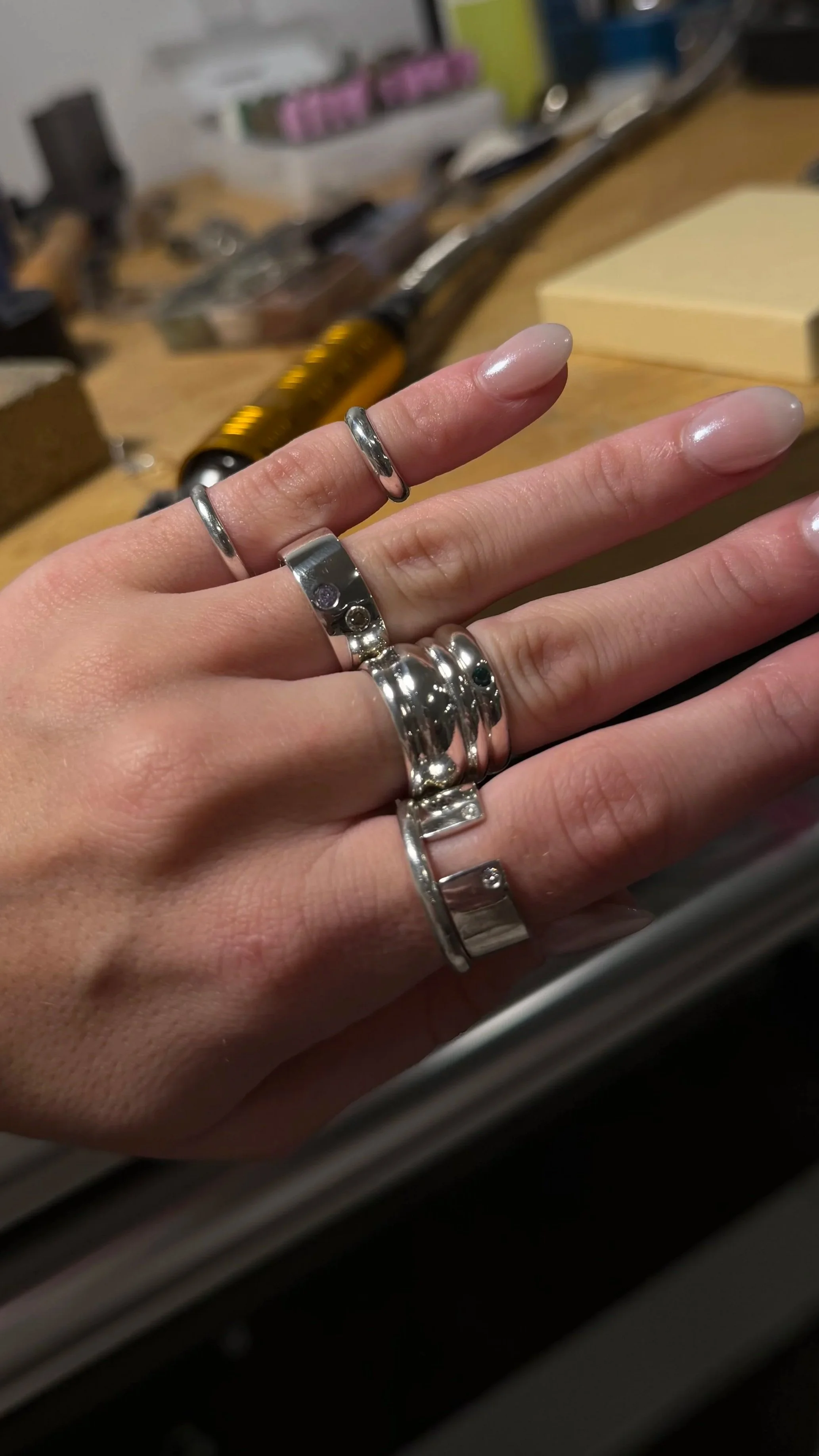 Close-up of a hand wearing multiple silver rings, with a blurred workbench and tools in the background.