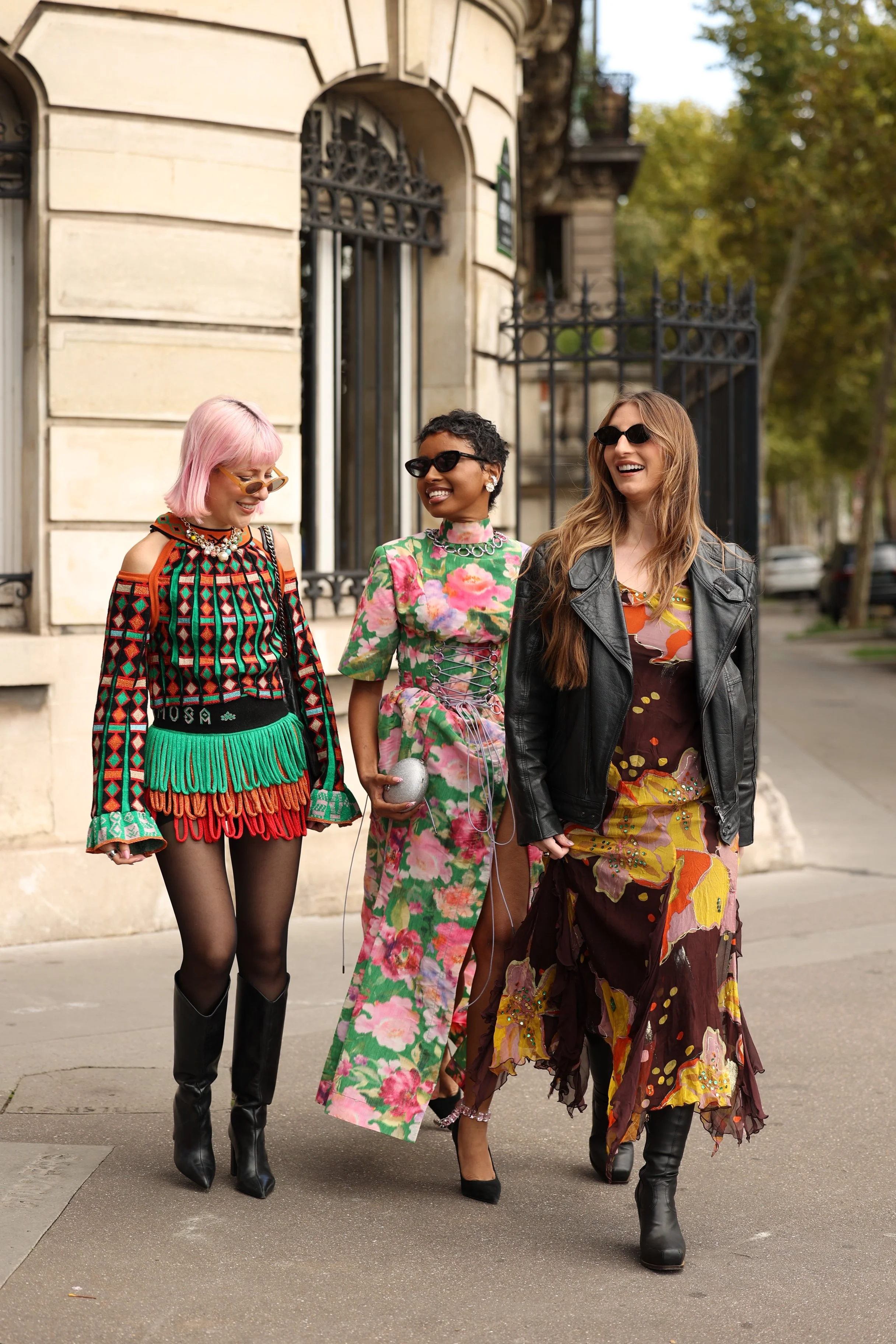 Quatro mulheres caminhando na rua, usando roupas coloridas e estilosas, com árvores ao fundo.
