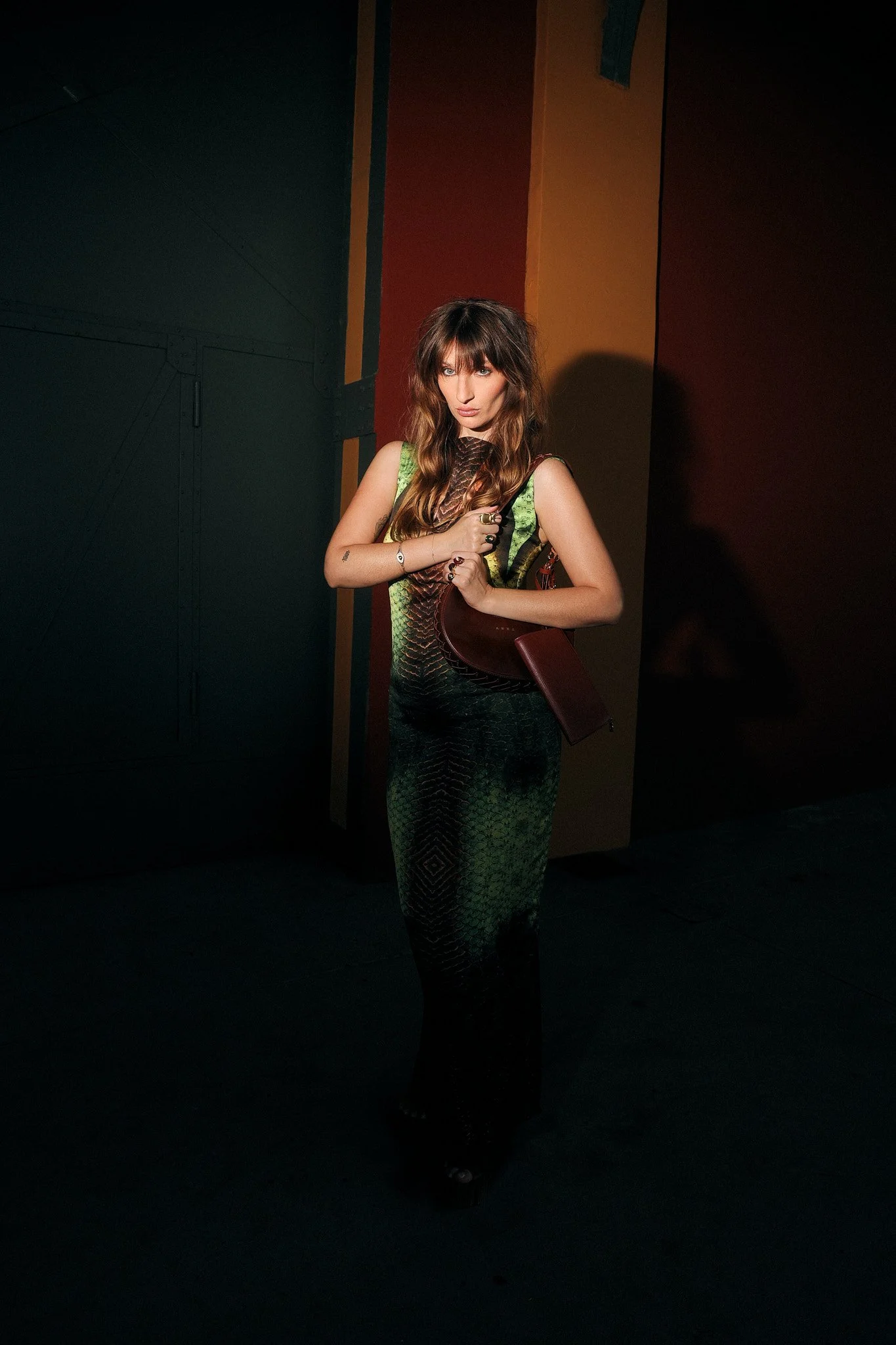 Mulher com cabelo castanho e vestido verde com detalhe em couro, posando com expressão séria em ambiente escuro com fundo colorido.