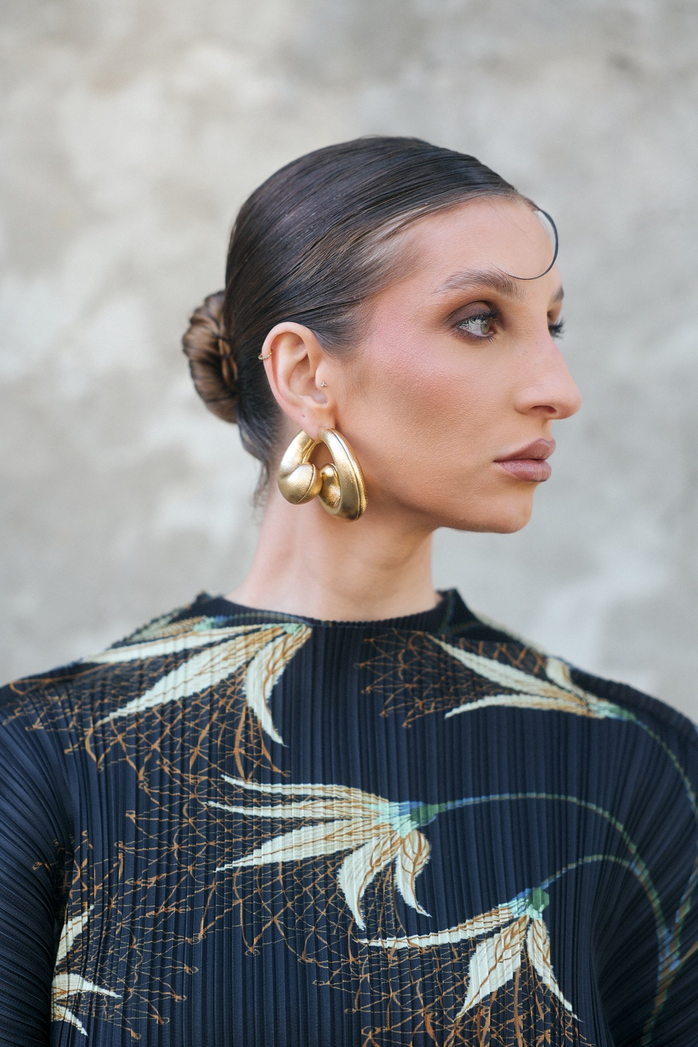 Retrato de uma mulher com cabelo escuro e preso em um coque. Ela usa brincos grandes dourados e uma blusa com estampa de cercas de ferro, em fundo neutro.