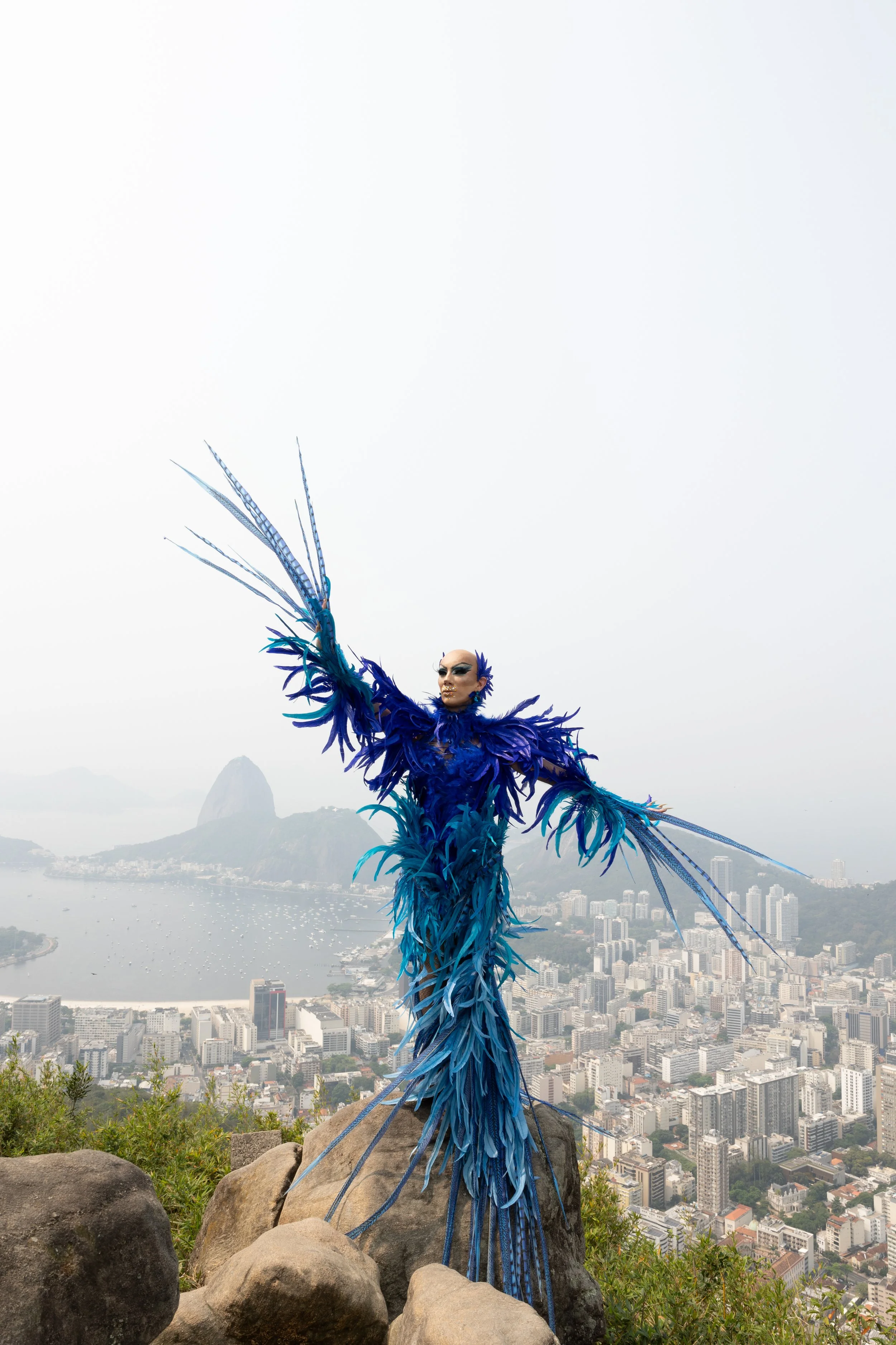 Figura de uma pessoa com fantasia azul de penas, posando com vista para a cidade e o mar ao fundo.