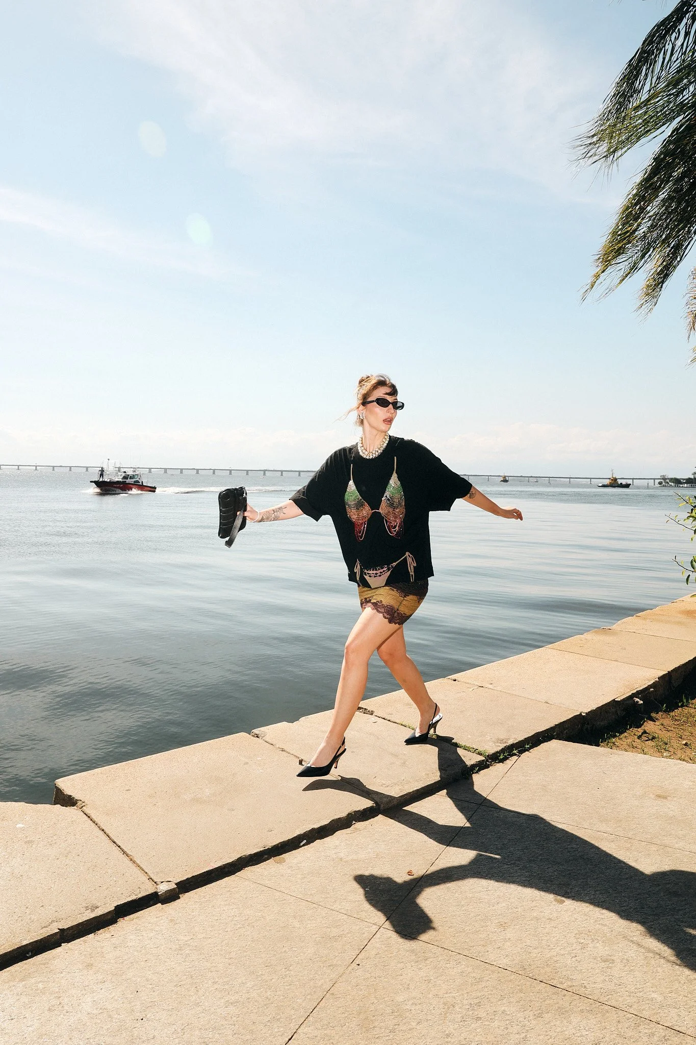 Mulher com cabelo preso, óculos escuros, camiseta preta com estampa de borboleta, saia de estampa colorida e salto alto, caminhando ao longo de um rio, com barcos ao fundo, sob céu claro, ao lado de um coqueiro