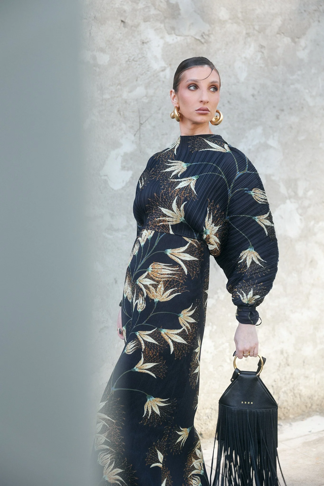 Mulher com cabelo preso, usando vestido longo com estampa de folhas e flores em tons de preto, branco e dourado, segurando uma bolsa de mão preta com detalhes dourados e franjas.