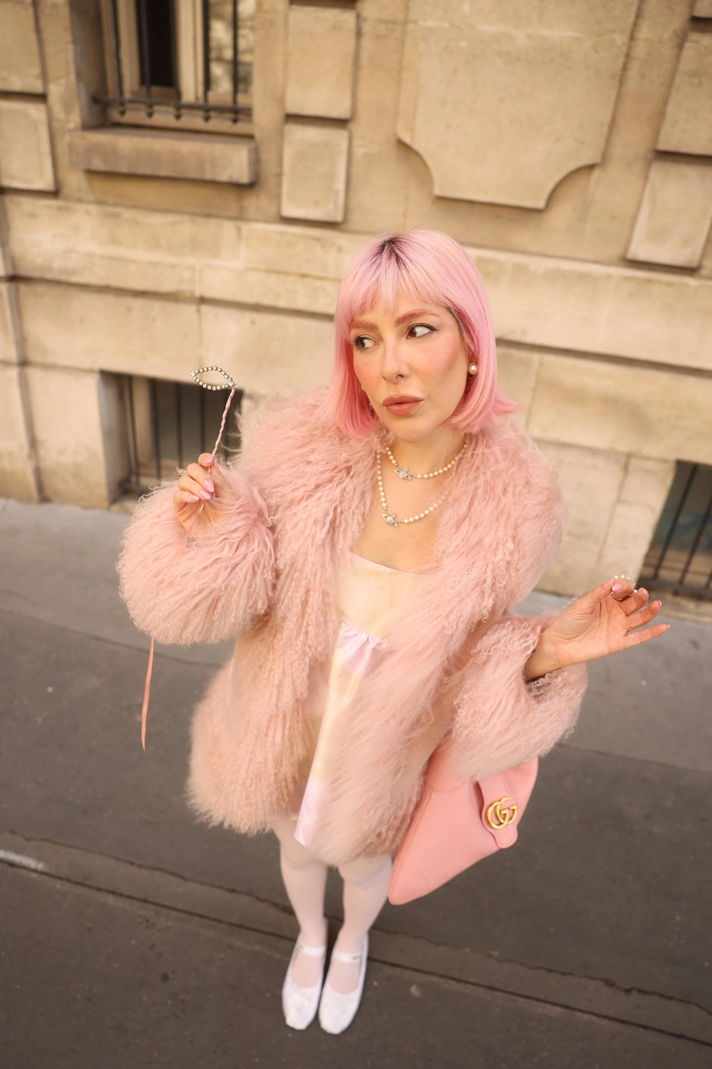Mulher com cabelo rosa, usando vestido rosa, casaco rosa de pelúcia, acessórios de pérolas, segurando uma máscara de maquiagem, na rua em frente a um edifício de pedra, vista de cima para baixo.