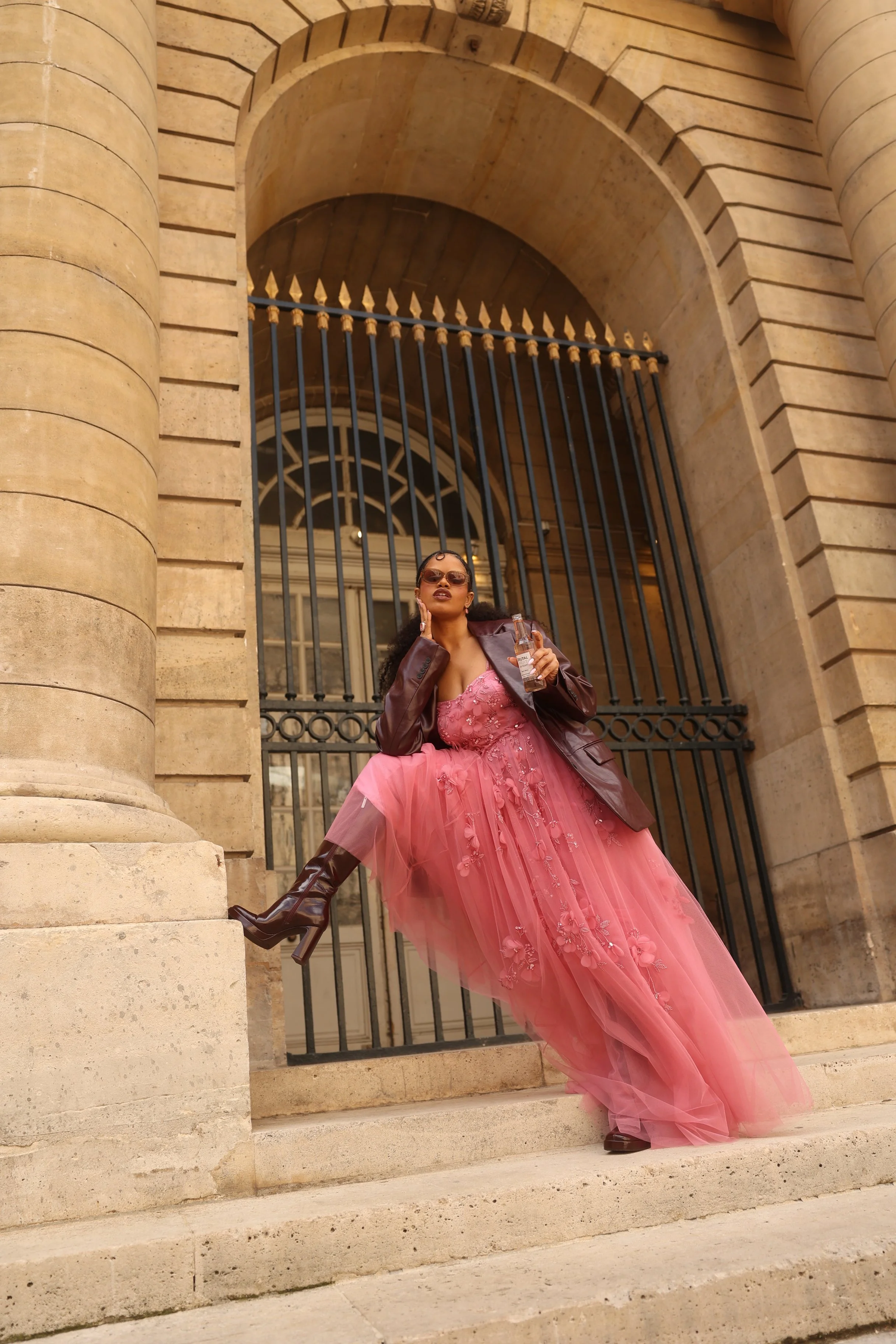 Mulher com vestido rosa longo de tule, jaqueta marrom, óculos escuros, sapatos de salto alto de couro marrom, posando de um lado de uma escadaria de pedra com estrutura de ferro preto e atrás uma porta de vidro grande com moldura branca, em frente a 