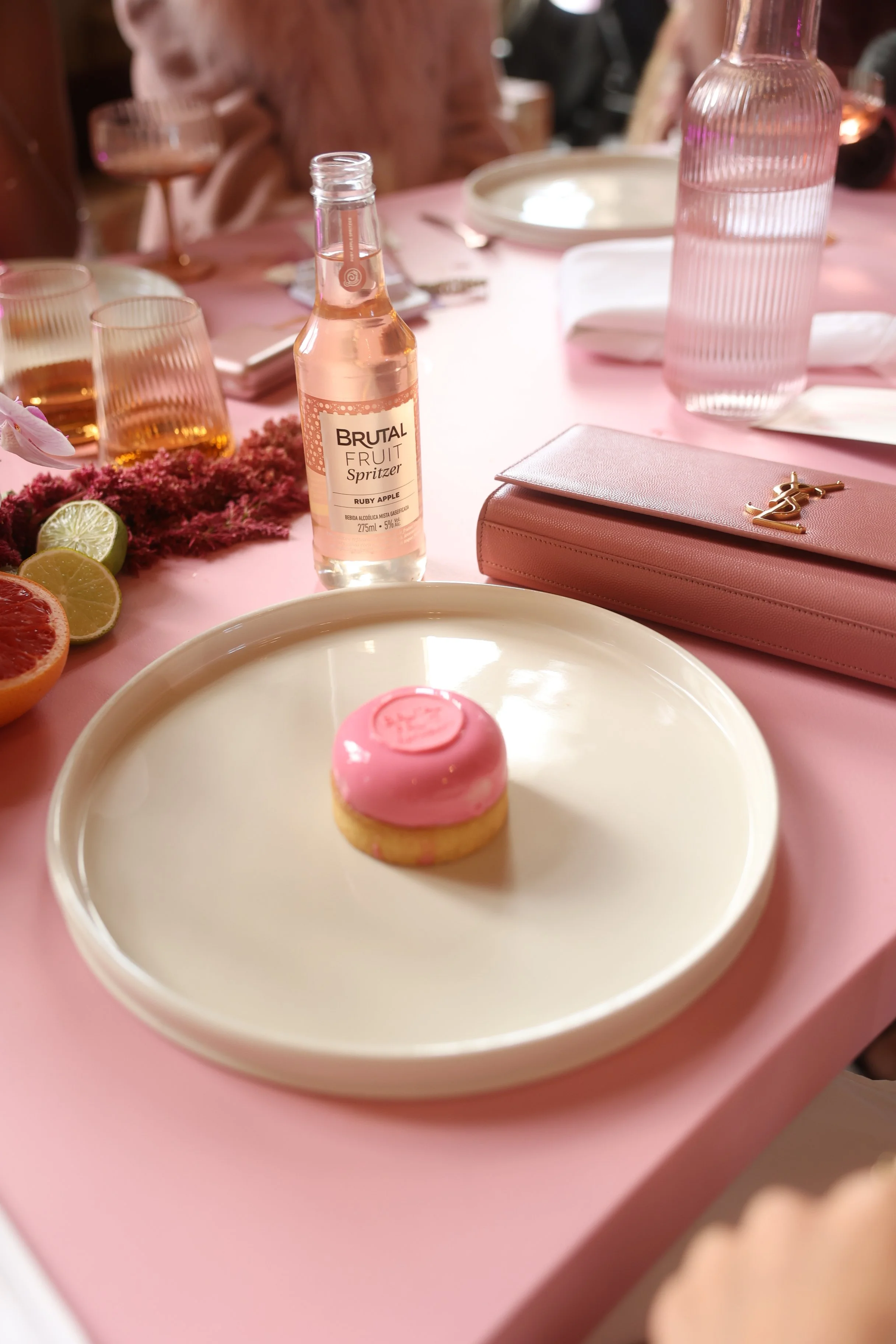 Mesa decorada com rosa e detalhes dourados, com uma garrafinha de suco de maçã, frutas cortadas, uma caixa de maquiagem rosa e uma taça ao fundo.